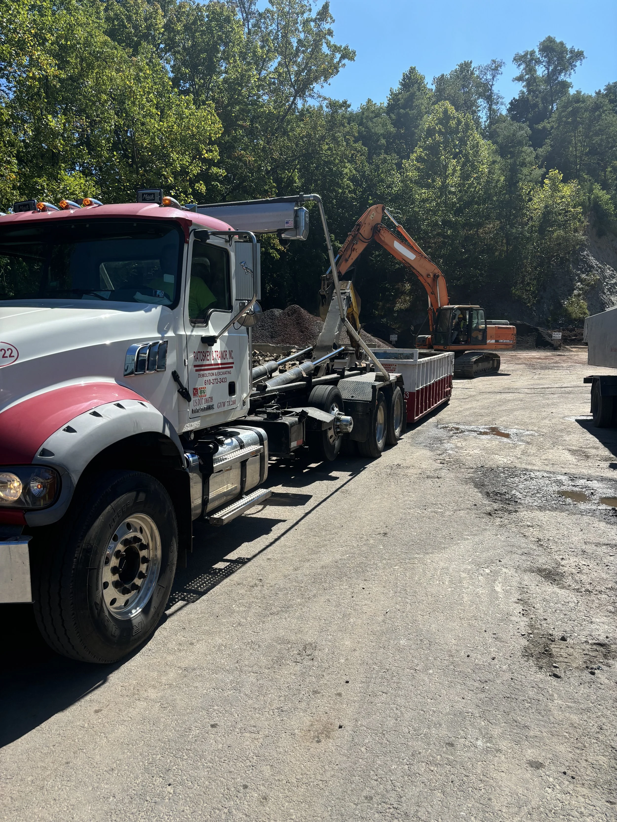 Construction site with a white dump truck and an orange excavator near a hillside with dense trees.