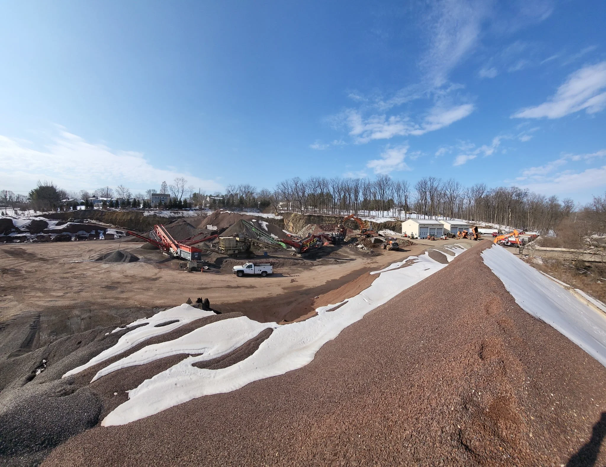 Construction site with piles of gravel and snow, heavy machinery, and storage buildings under a partly cloudy sky.