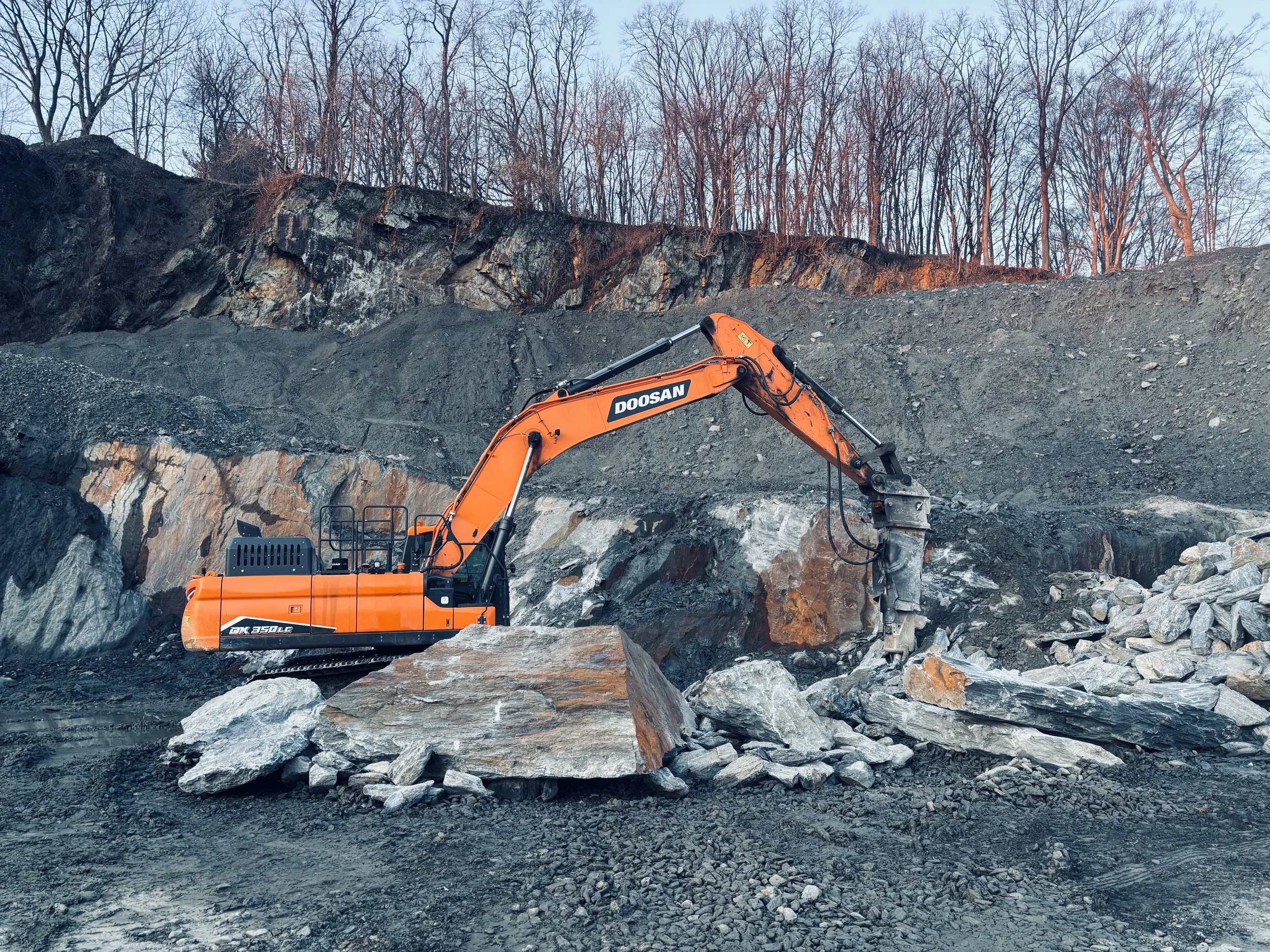 An orange Doosan excavator working on a rocky outdoor construction site, breaking large rocks into smaller pieces with a rocky hillside and leafless trees in the background.