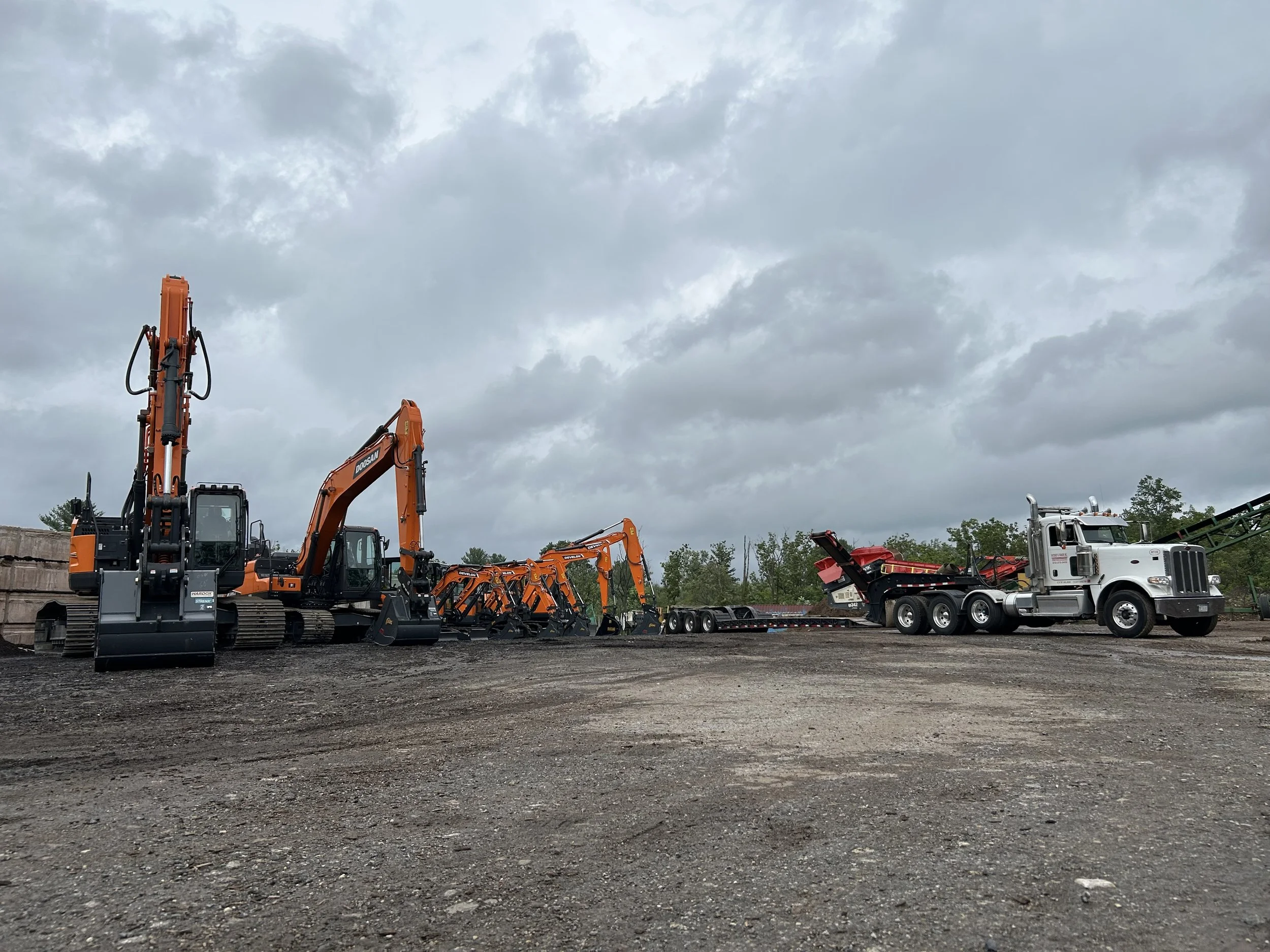 Construction equipment including excavators and a dump truck parked on a gravel lot with cloudy sky overhead.