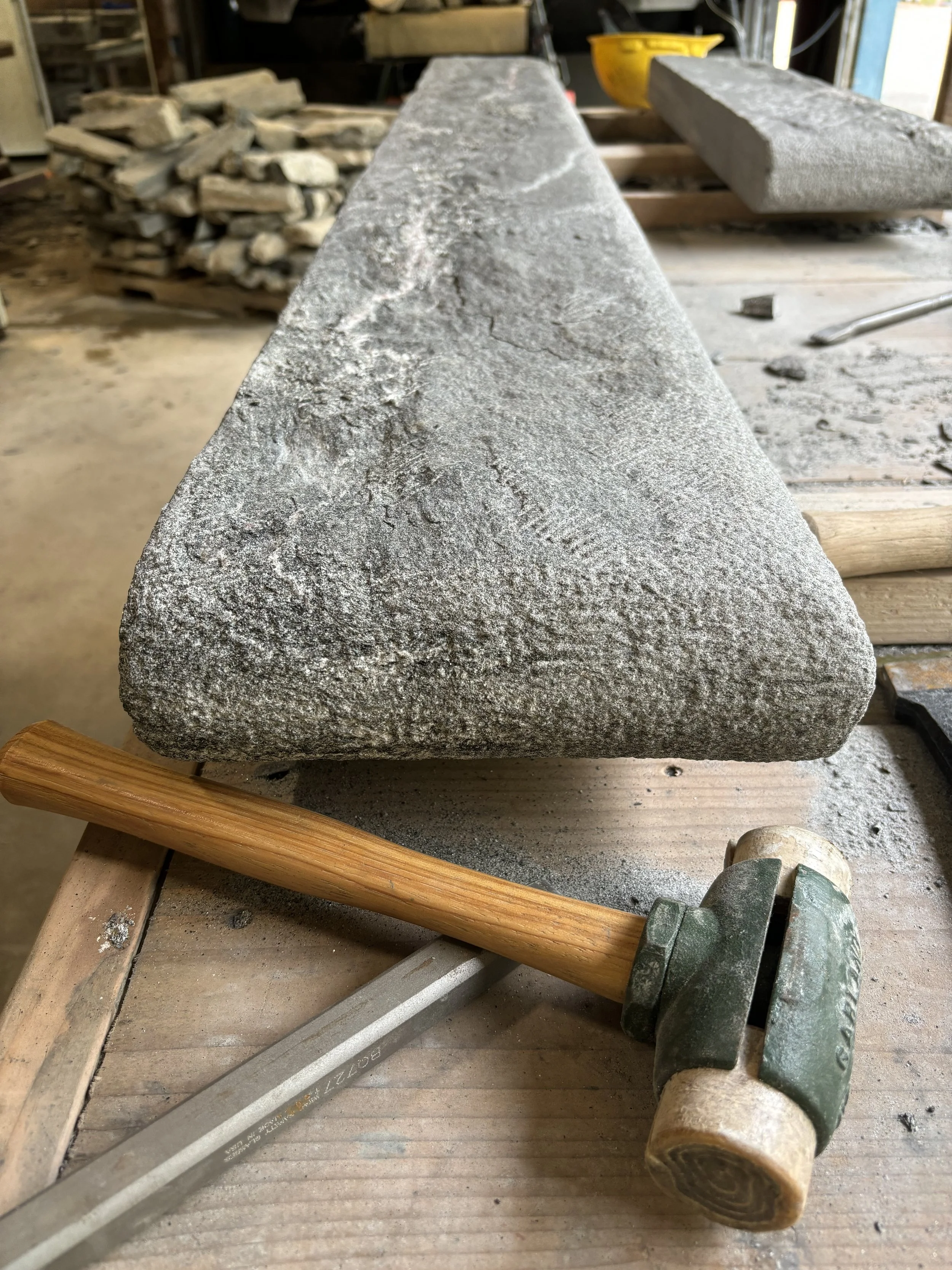 A large, rough-textured stone in a workshop with a hammer with a wooden handle and a metal chisel resting on a wooden work surface.