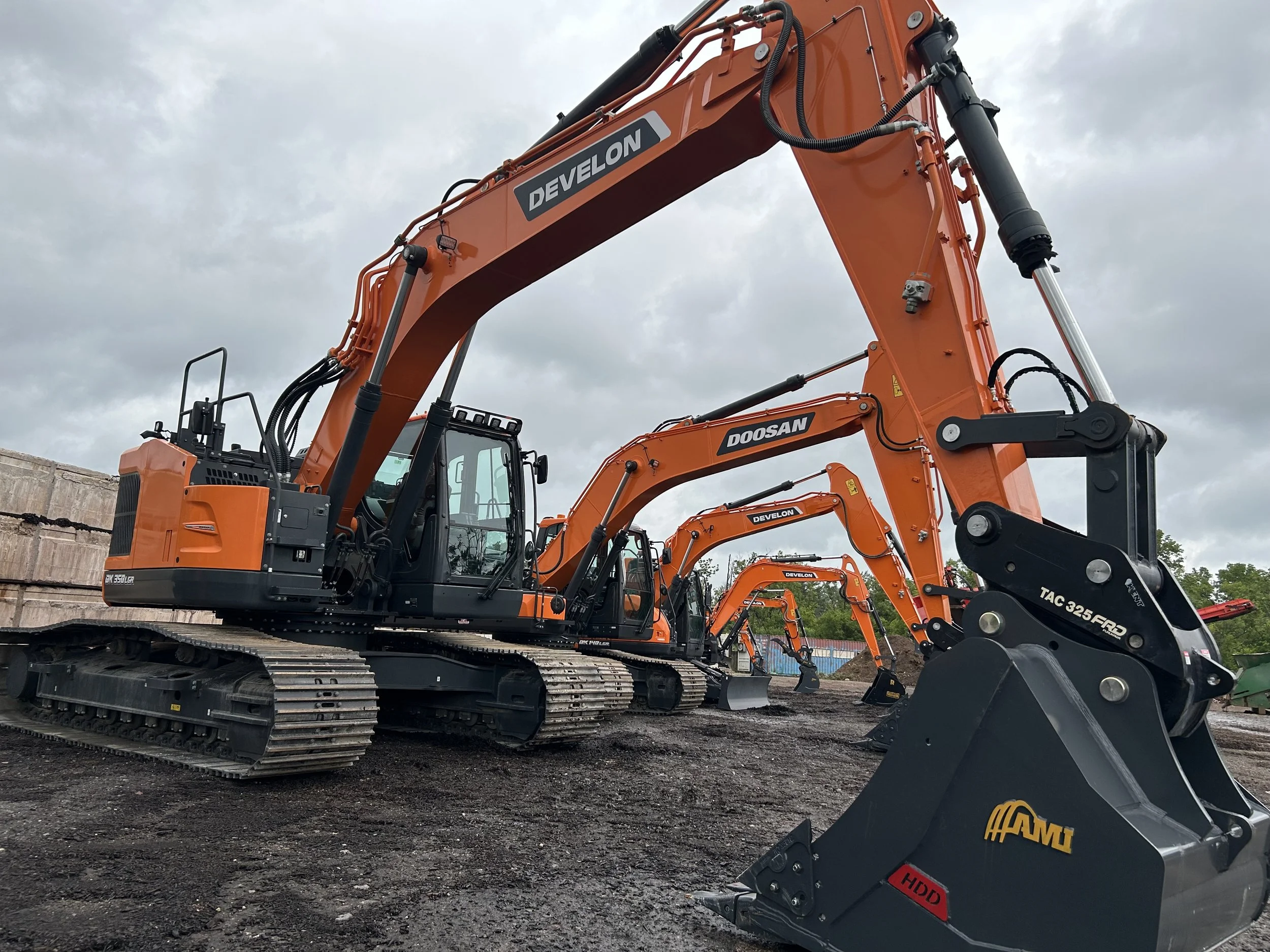 Several orange and black excavators lined up on a dirt construction site under a cloudy sky.