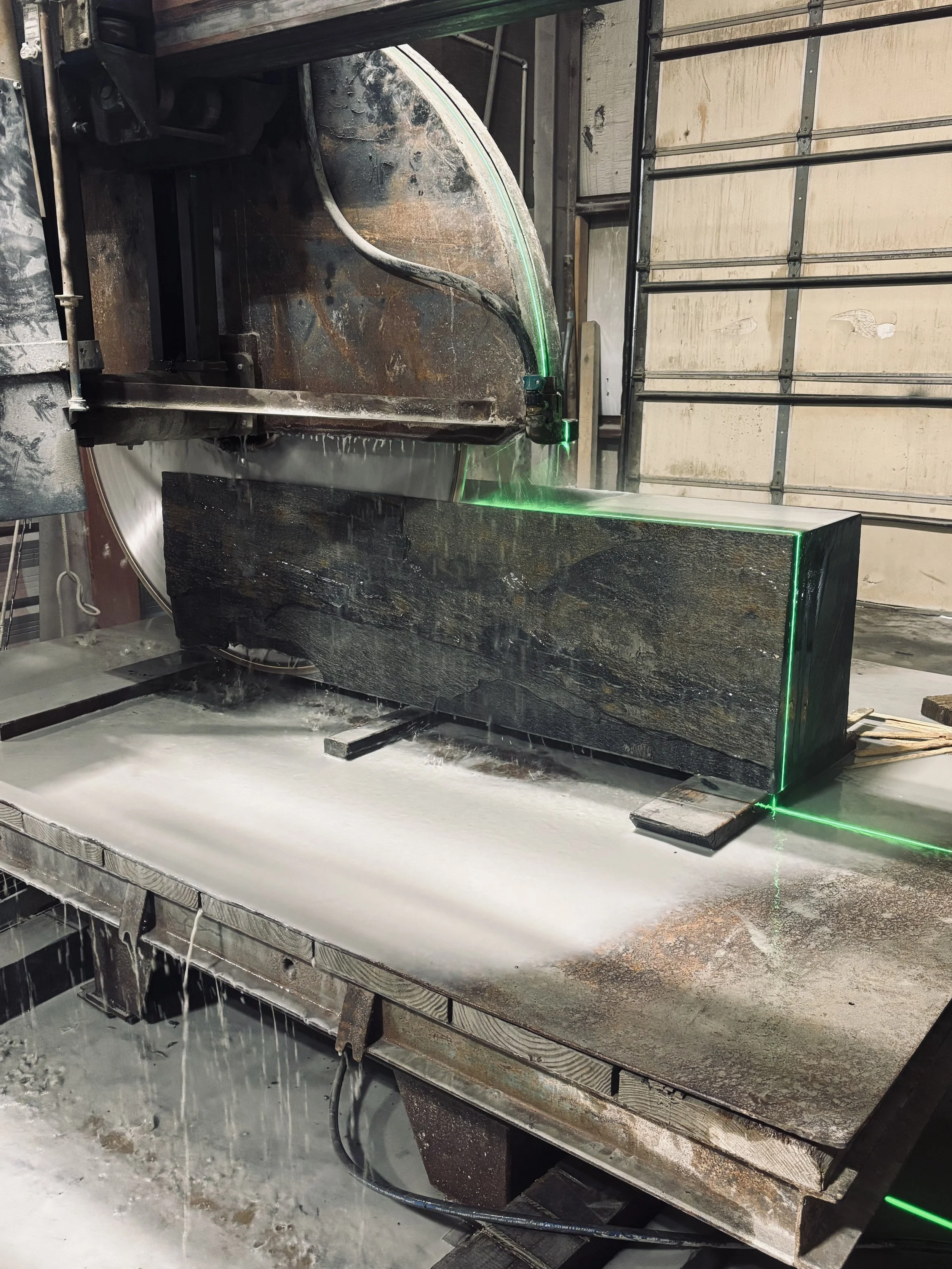 A large industrial saw cutting through a dark stone slab in a workshop, with water used for cooling, and green laser guides outlining the cut.