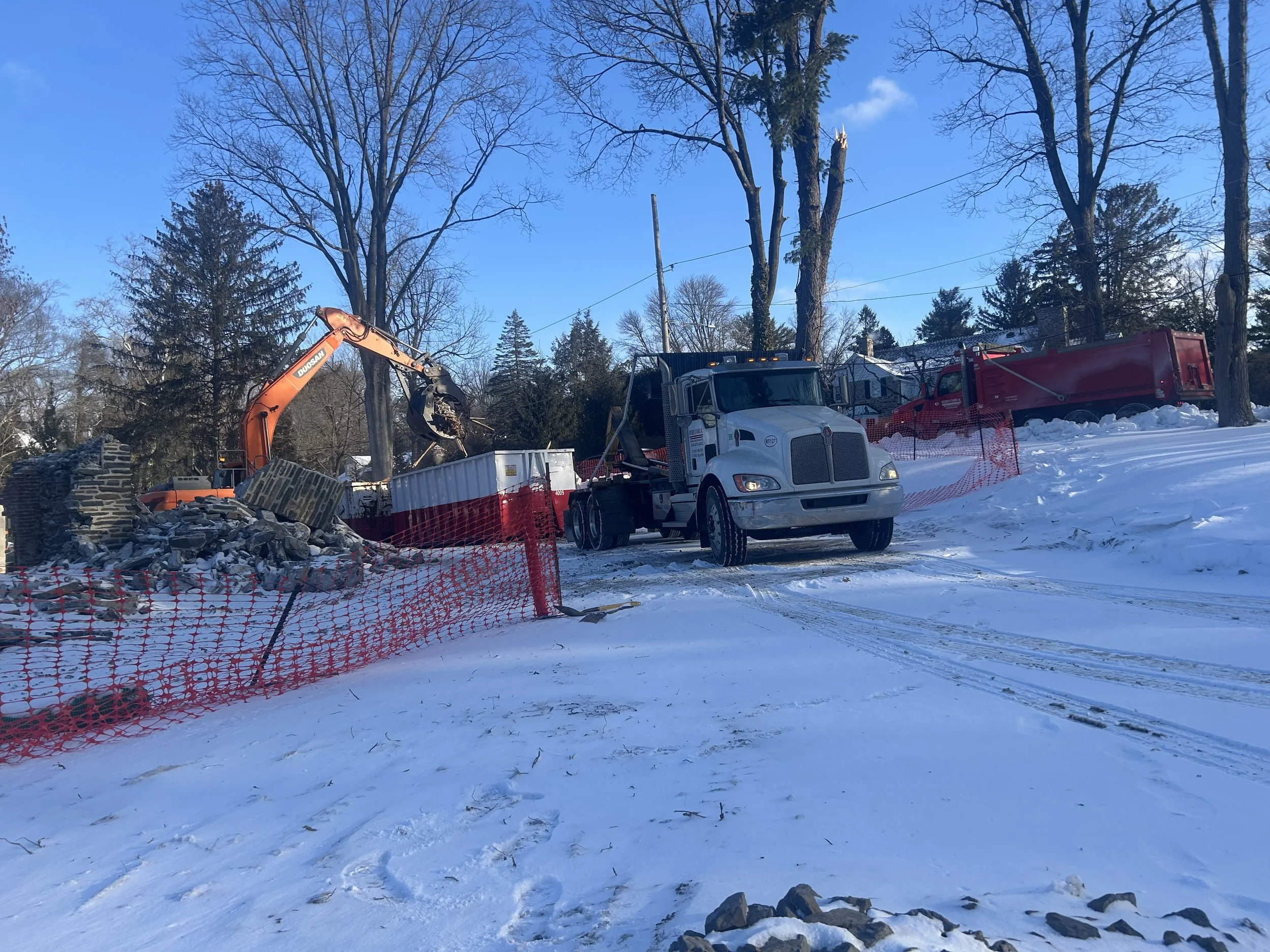 Construction site in snowy area with excavator removing debris, surrounded by orange safety fencing, vehicles, leafless trees, and snow-covered ground.