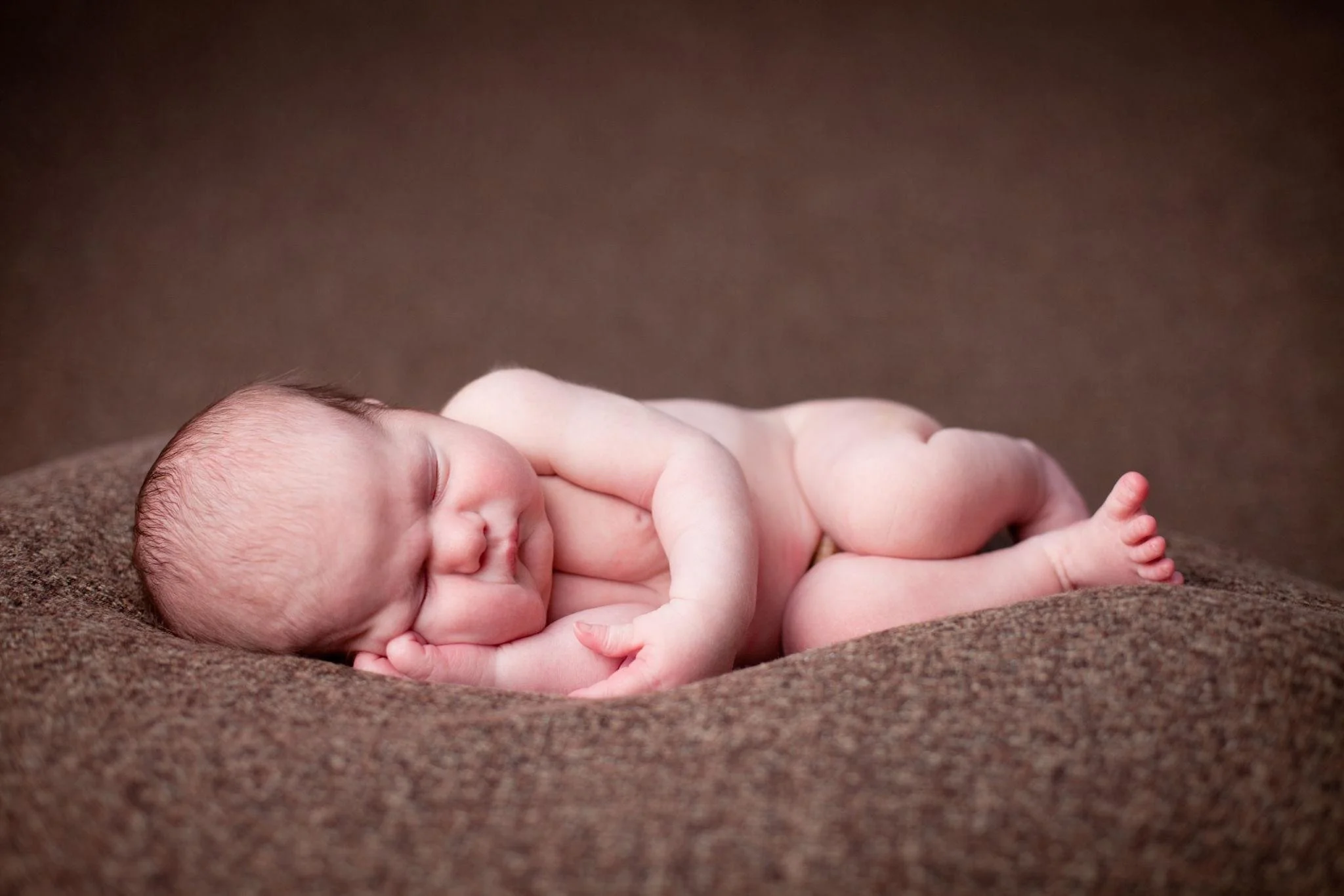 A sleeping newborn baby lying on a brown blanket with hands tucked under its face.