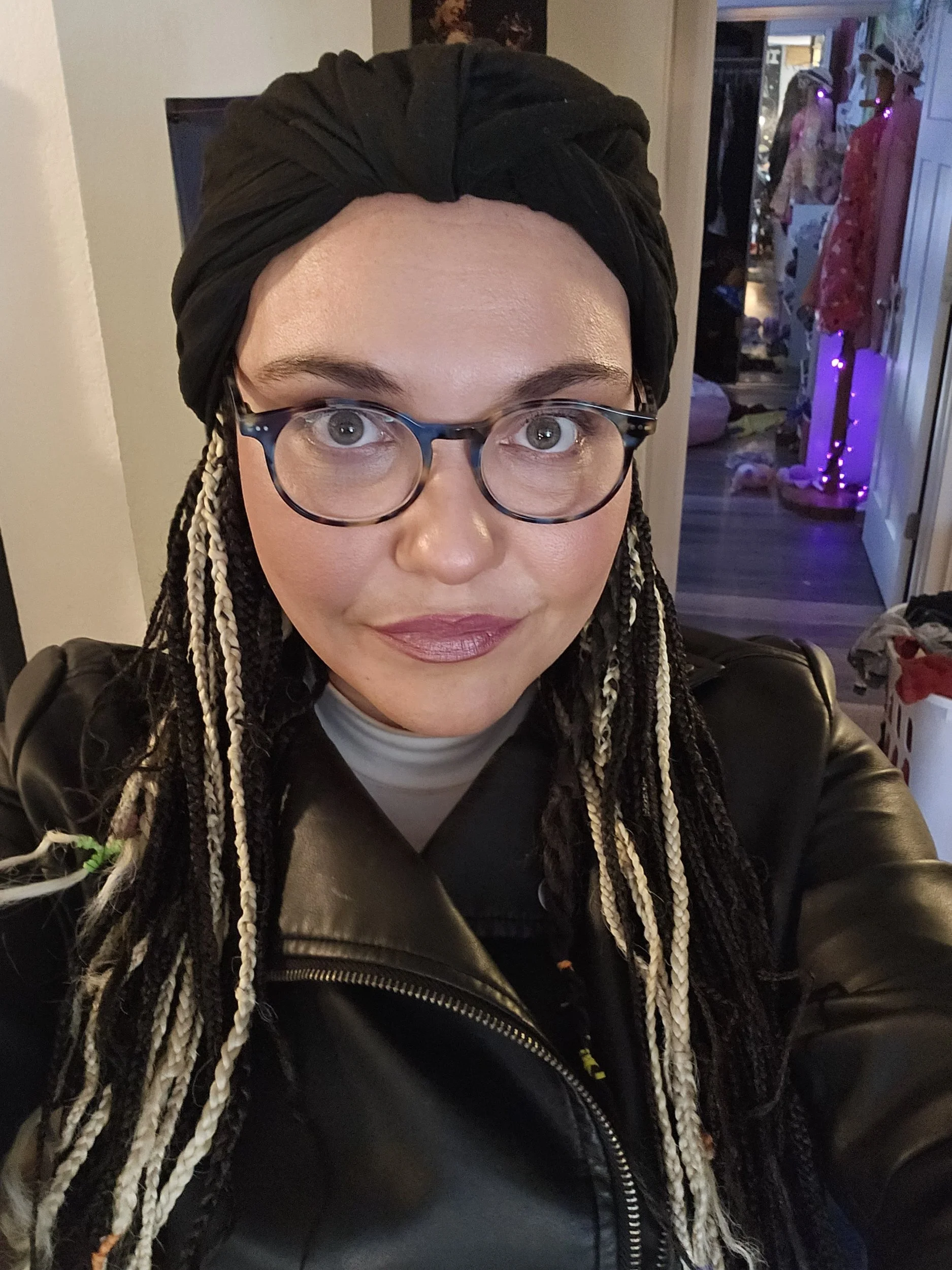 A woman with glasses, braided hair with blonde streaks, wearing a black headscarf, black leather jacket, and lipstick, taking a selfie indoors with a decorated Christmas tree and festive decor in the background.