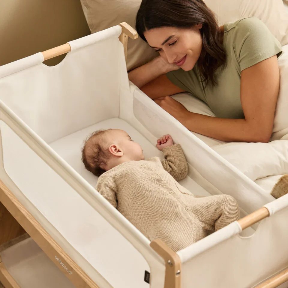 A woman lying on a bed, smiling gently while looking at a sleeping baby in a white bassinet.