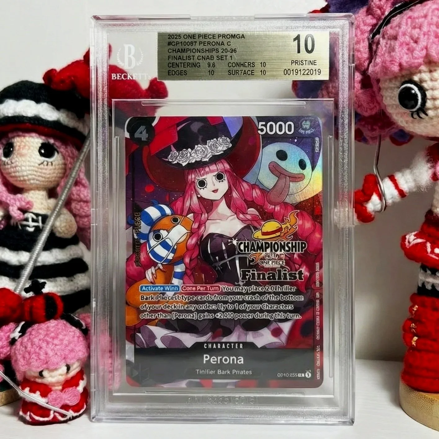 Close-up of a graded collectible card encapsulated in a plastic case, featuring anime-style character Perona with pink hair and black outfit, surrounded by colorful characters and symbols from the One Piece series, with grading information and text indicating it is a Finalist card.