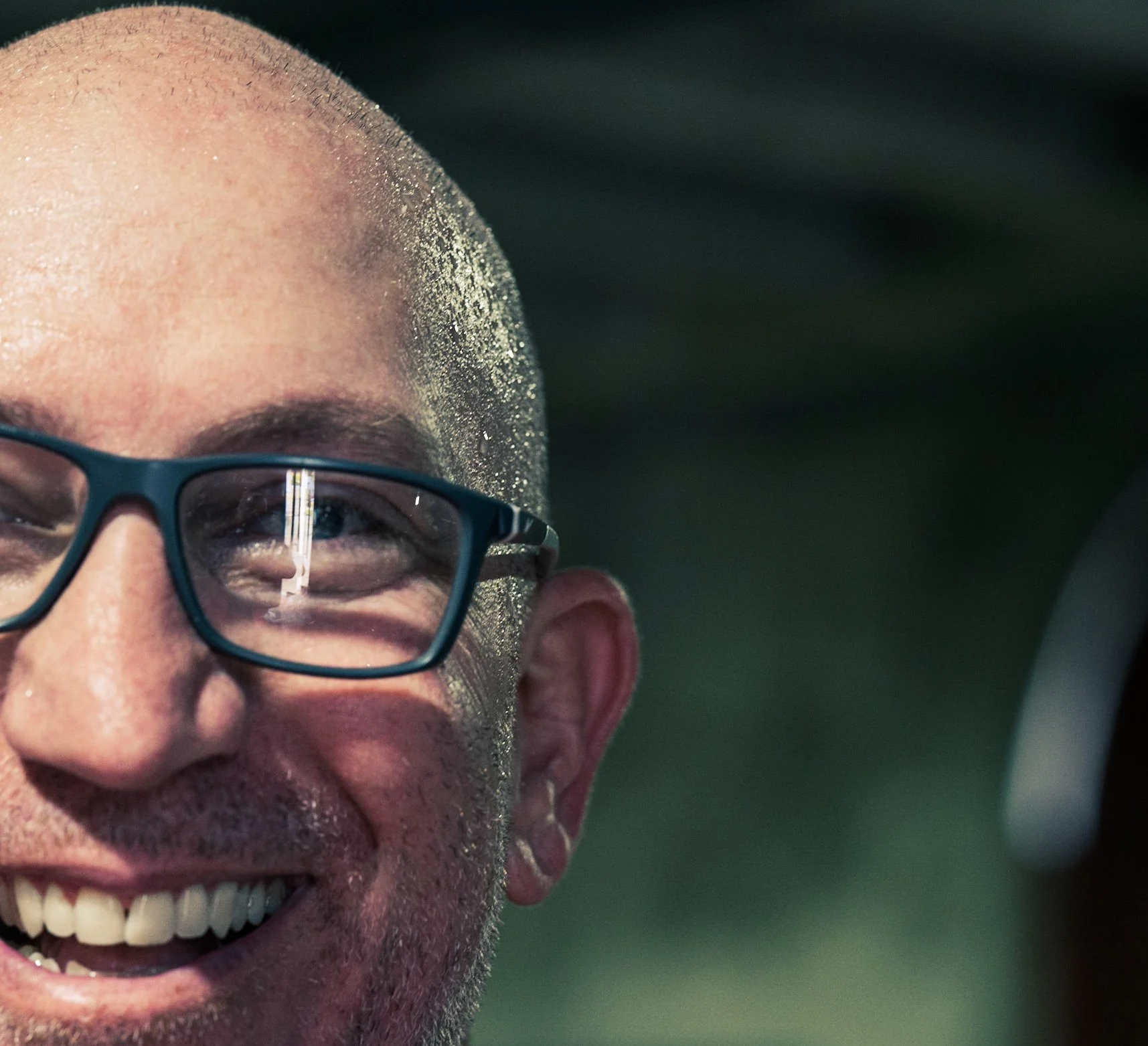 Close-up of a bald man with glasses smiling, with sunlight reflecting off his face and hair, and a dark green blurred background.