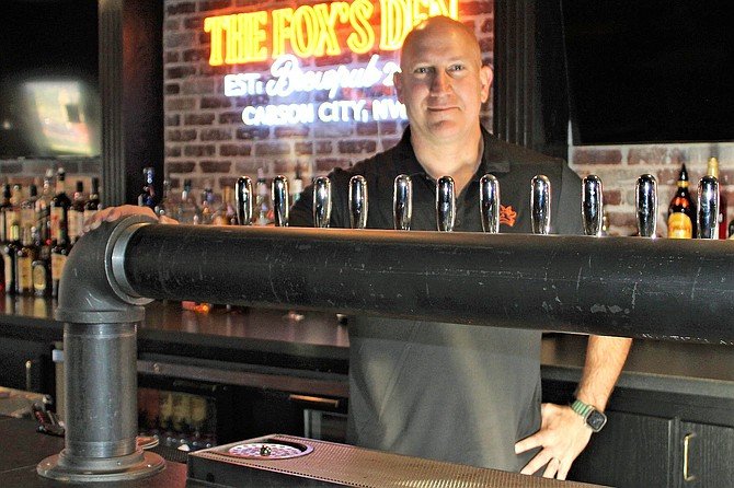 A bartender standing behind a bar with beer taps on a black pipe faucet system, inside a bar named 'The Fox's Den' with a neon sign in the background.
