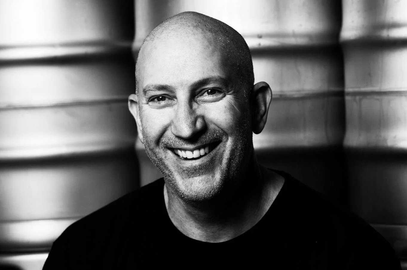 A smiling man with a shaved head and stubble, wearing a black shirt, stands in front of what appears to be large metal barrels in a black-and-white photograph.