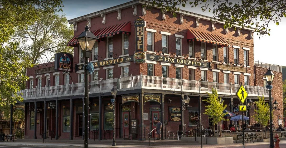 A three-story red brick building housing The Fox Brewpub and Hotel, with outdoor seating, street lamps, and a pedestrian crossing sign in front, surrounded by trees.