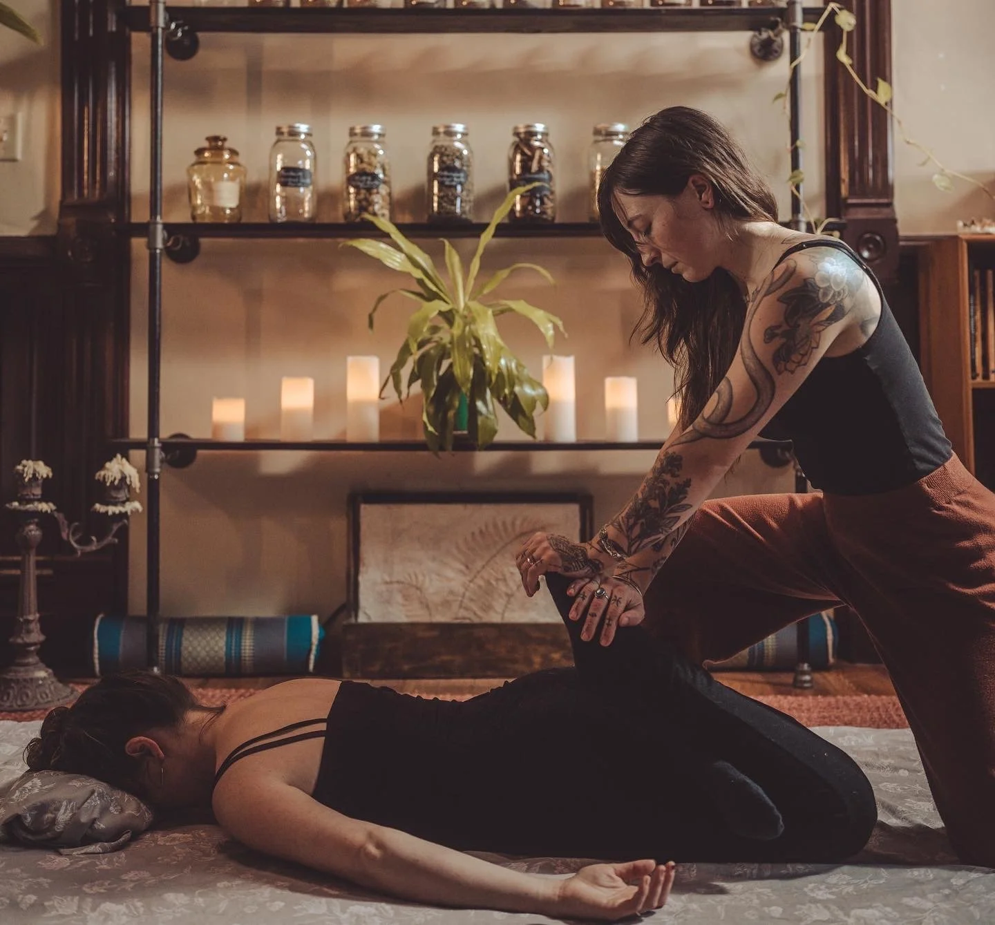 A woman with tattoos practicing yoga on the floor while another woman assists her in a warm, cozy room decorated with candles, plants, and shelves.