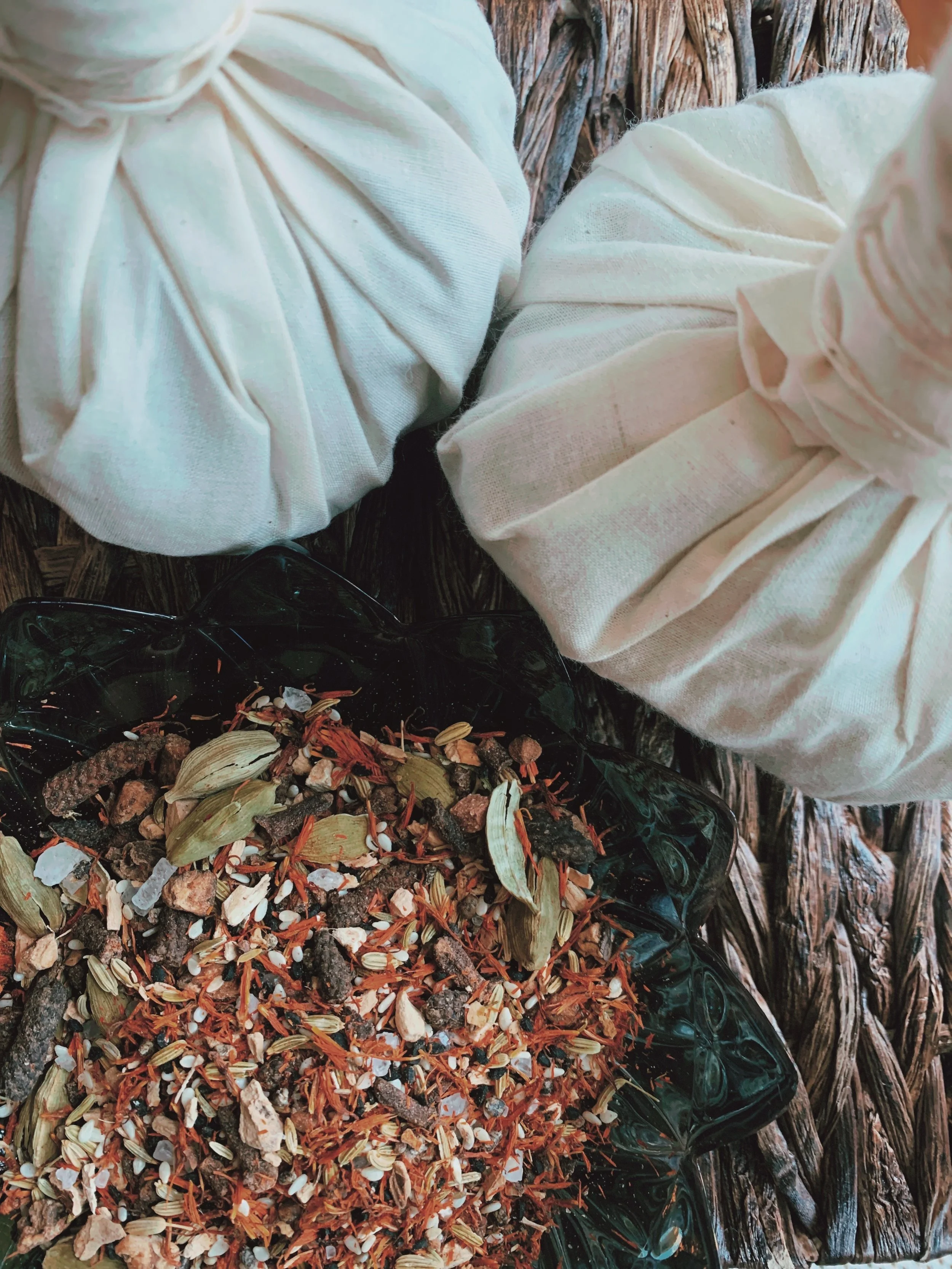 A black glass dish containing coffee grounds, dried flower petals, seeds, and small stones, placed on a wooden surface with two cream-colored cloth bundles nearby.