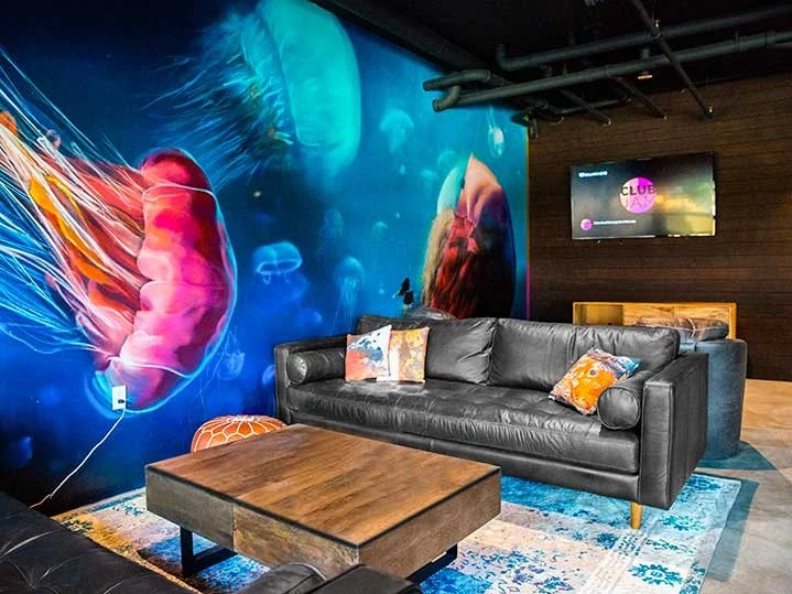 Modern lounge area with a large black leather sofa, four colorful throw pillows, a wooden coffee table, a vibrant underwater-themed mural of jellyfish on the wall, and a small wall-mounted television.