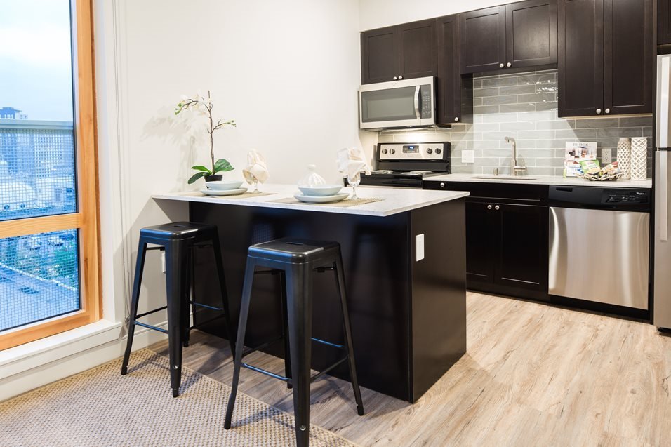 Modern kitchen with black cabinets, gray backsplash, stainless steel appliances, a small island with two black barstools, and a window overlooking a cityscape.