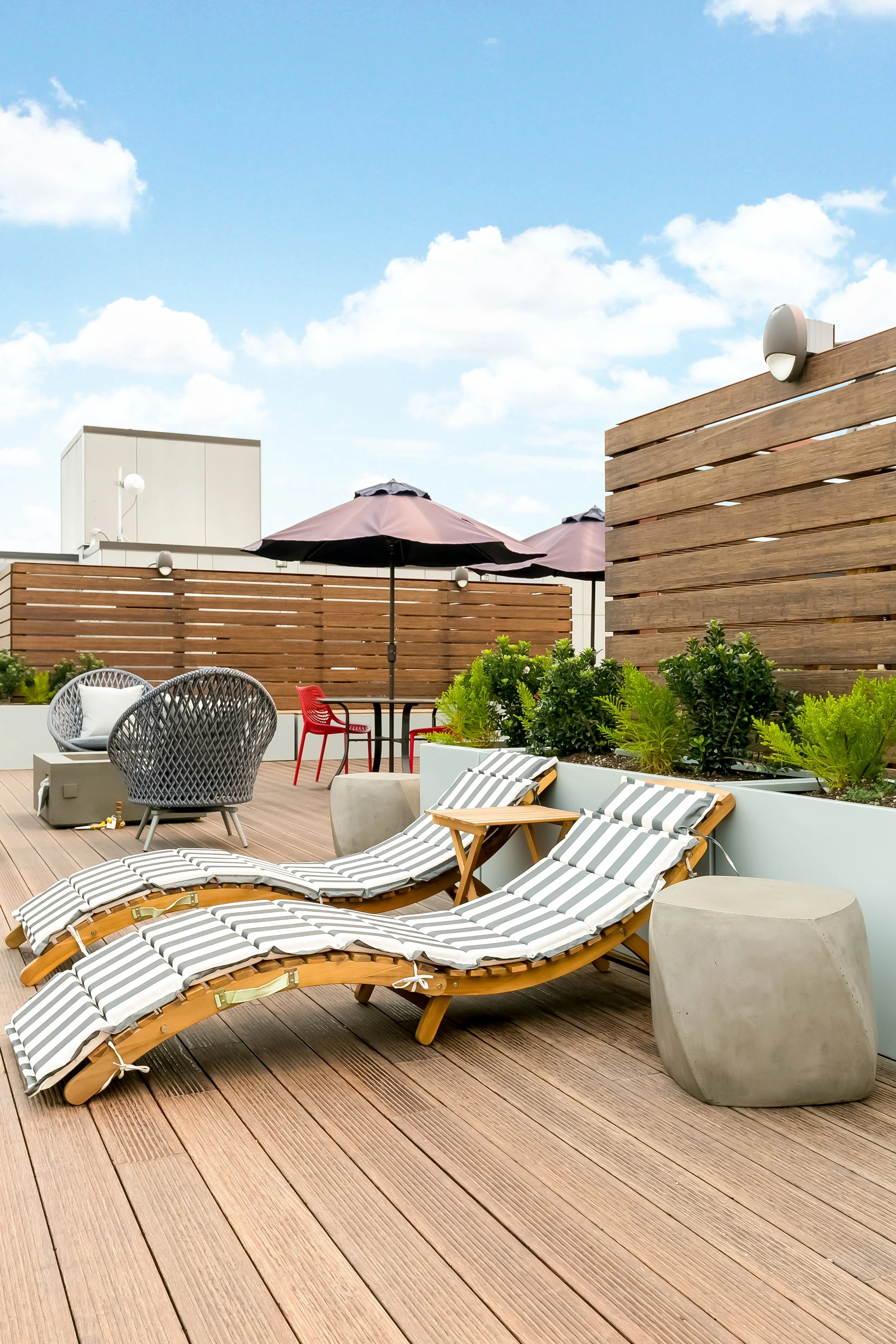 Rooftop terrace with outdoor furniture, striped lounge chairs, umbrellas, potted plants, and a wooden fence under a blue sky with clouds.