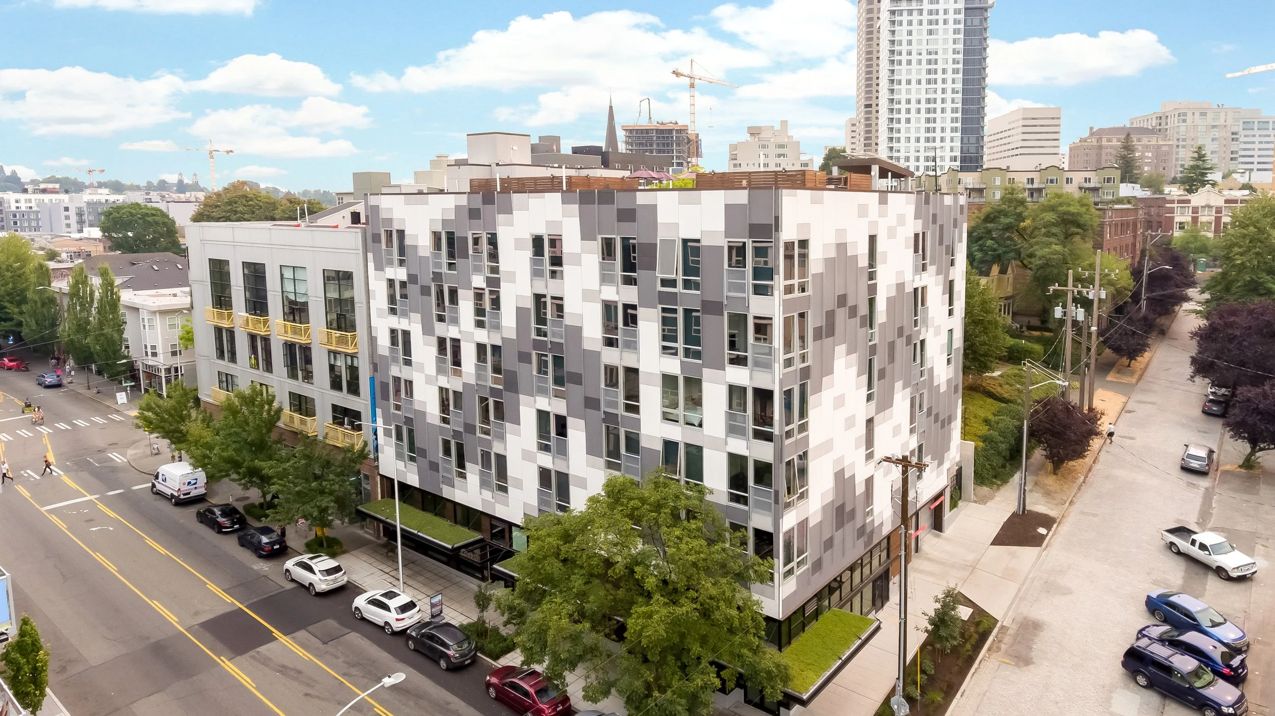 An aerial view of a modern multi-story building with a pixelated gray and white facade, surrounded by trees, parked cars, and streets in an urban area under a partly cloudy sky.