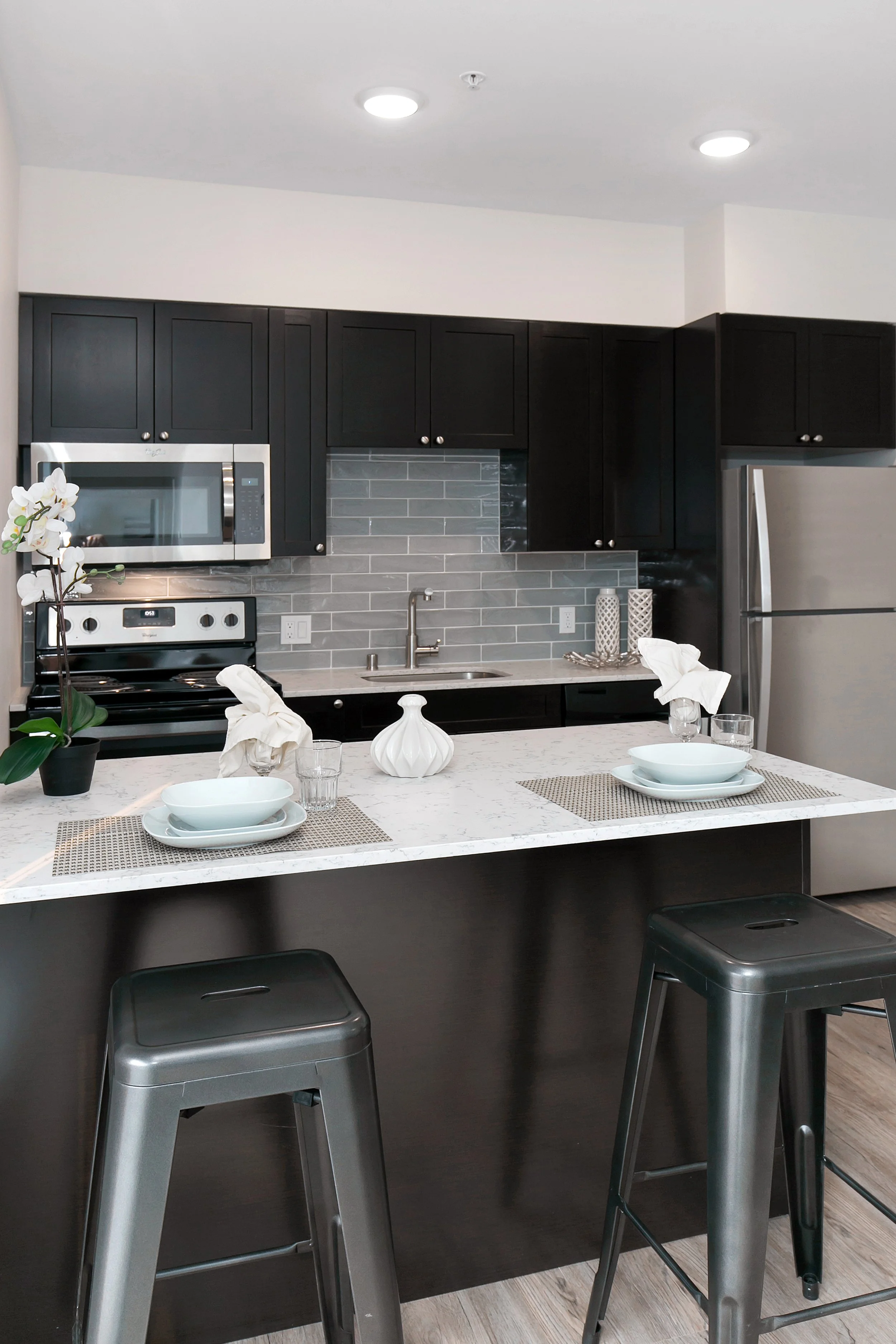 Modern kitchen with black cabinets, stainless steel refrigerator and microwave, gray tile backsplash, white countertop with place settings, and two metal bar stools.