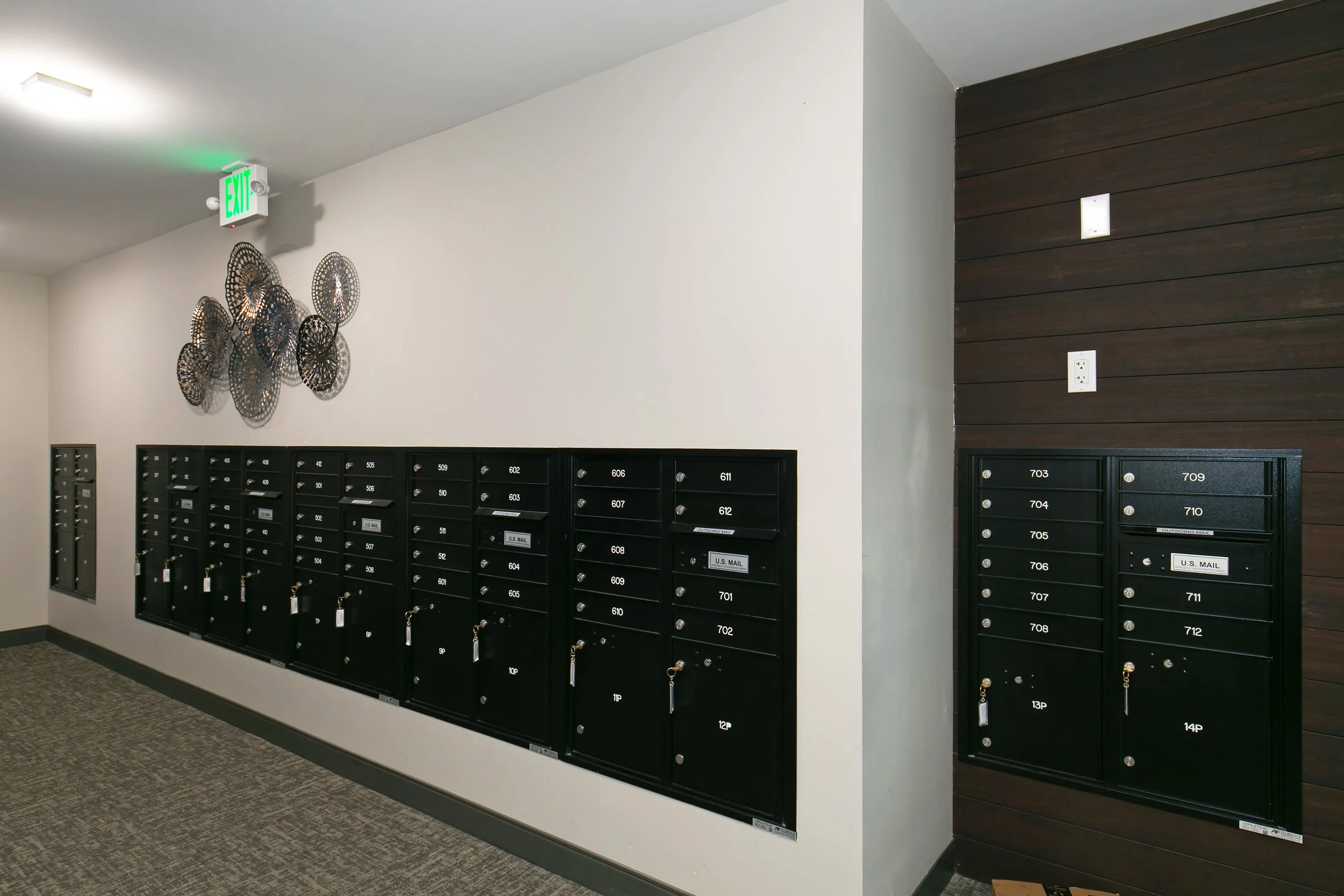 Indoor mailroom with black mailboxes mounted on a white wall, decorative metal wall art, and an emergency exit sign with green lettering.