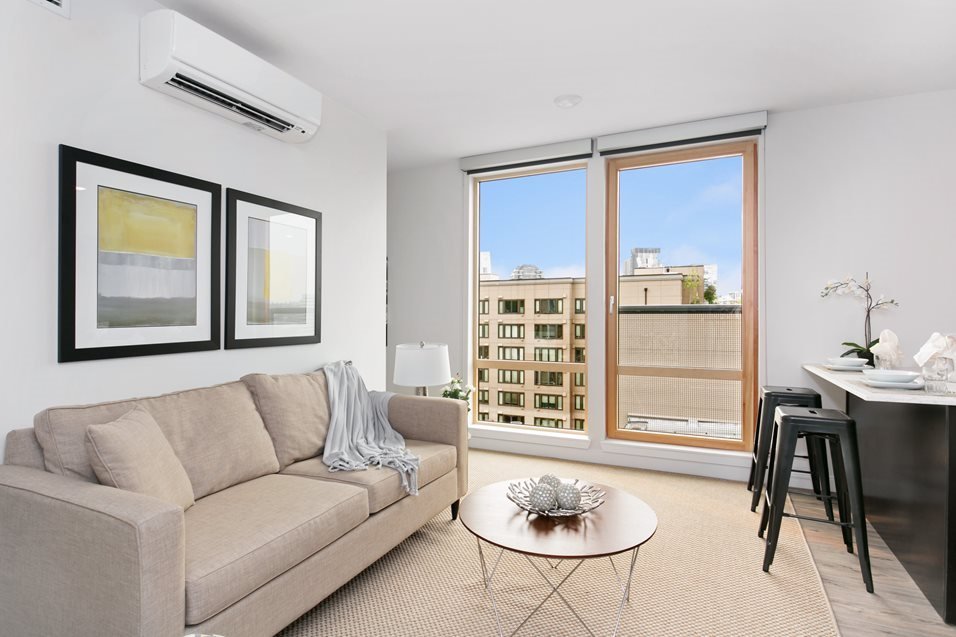 Living room with beige sofa, small round coffee table, wall art, window showing cityscape, air conditioning unit, side lamp, and kitchen bar with barstools.