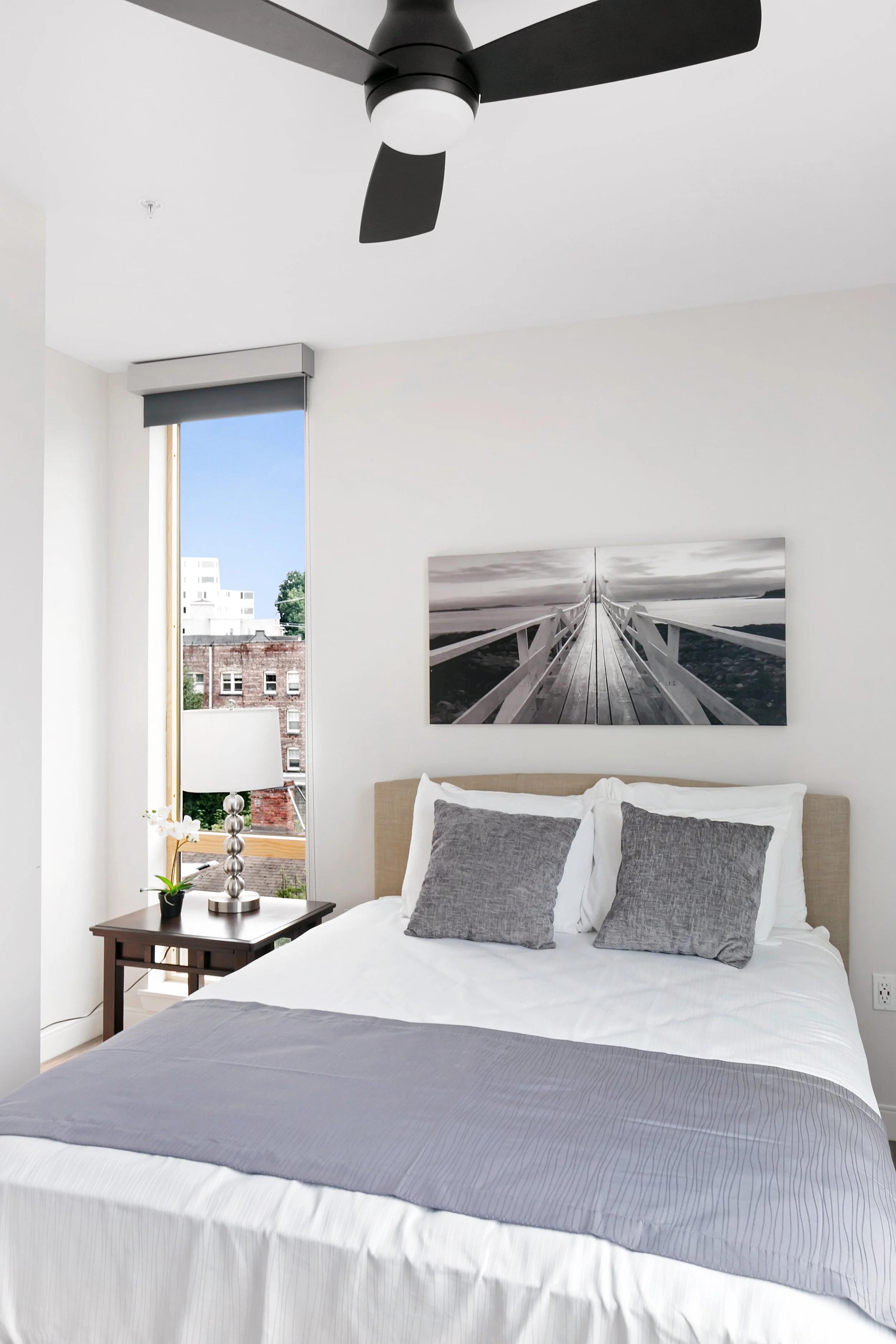 A modern bedroom with a neatly made bed, gray pillows, a wooden nightstand with a lamp and a small plant, a window with a city view, a black ceiling fan, and a black-and-white photograph of a pier on the wall.