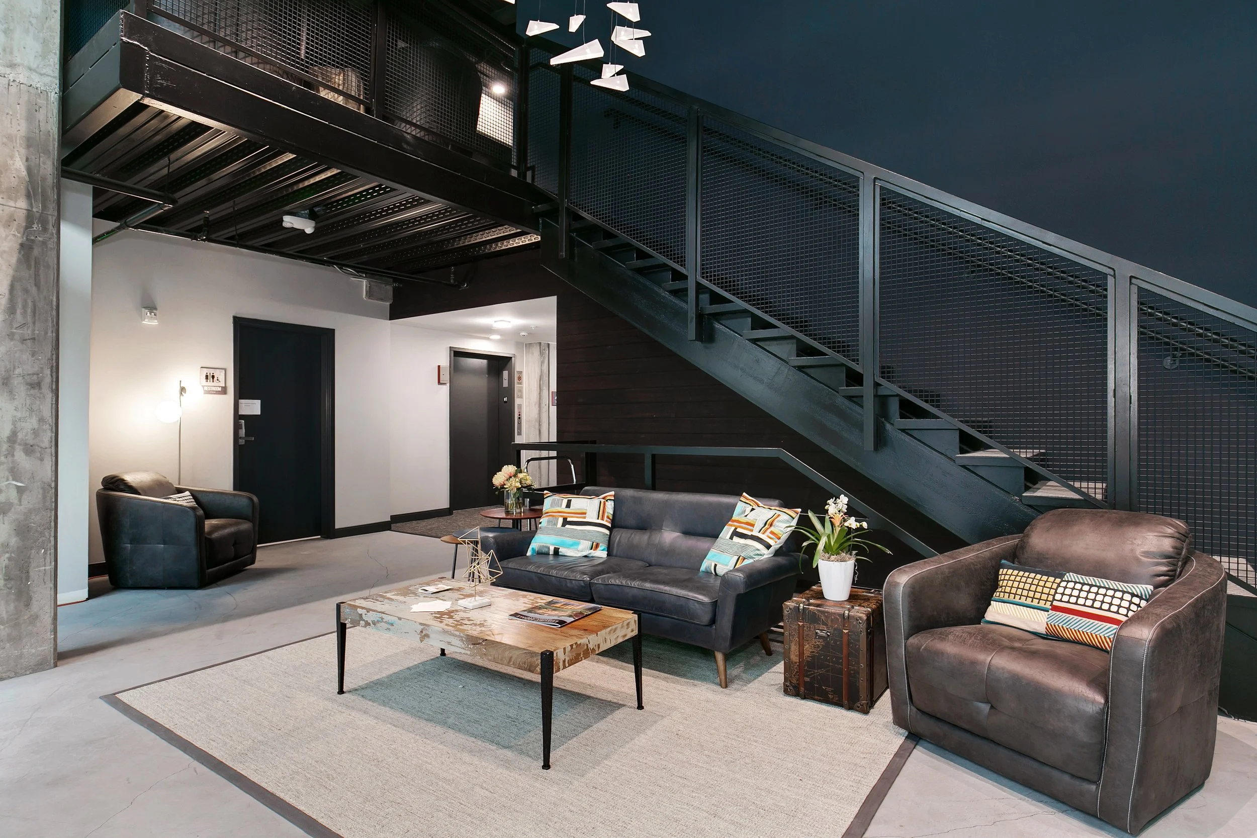 Modern lobby with black and brown leather armchairs, a sofa, a coffee table, and a rug. There are decorative pillows on the furniture, potted plants, and a staircase with black metal railings leading to a second floor.