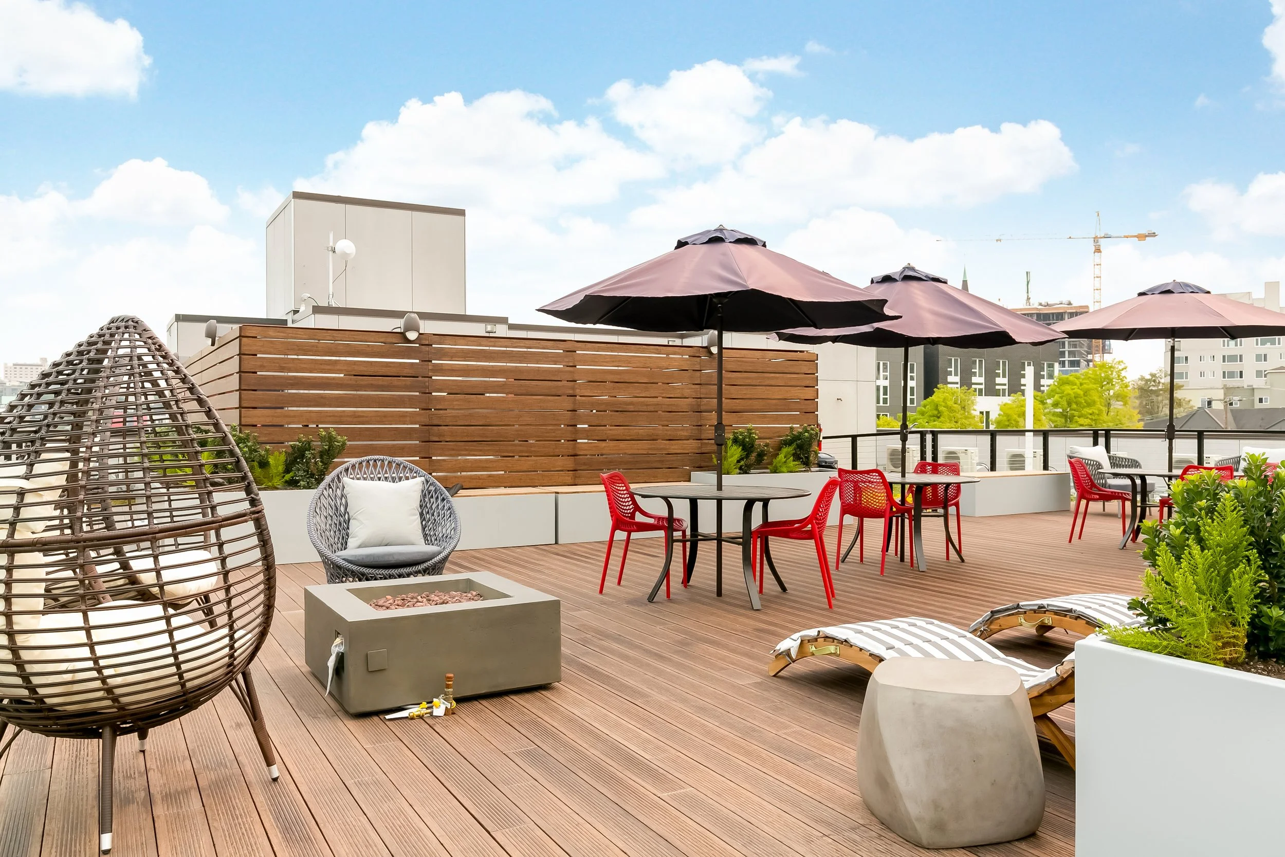 Rooftop patio with seating, umbrellas, a fire pit, lounge chairs, and potted greenery, overlooking a cityscape under a partly cloudy sky.