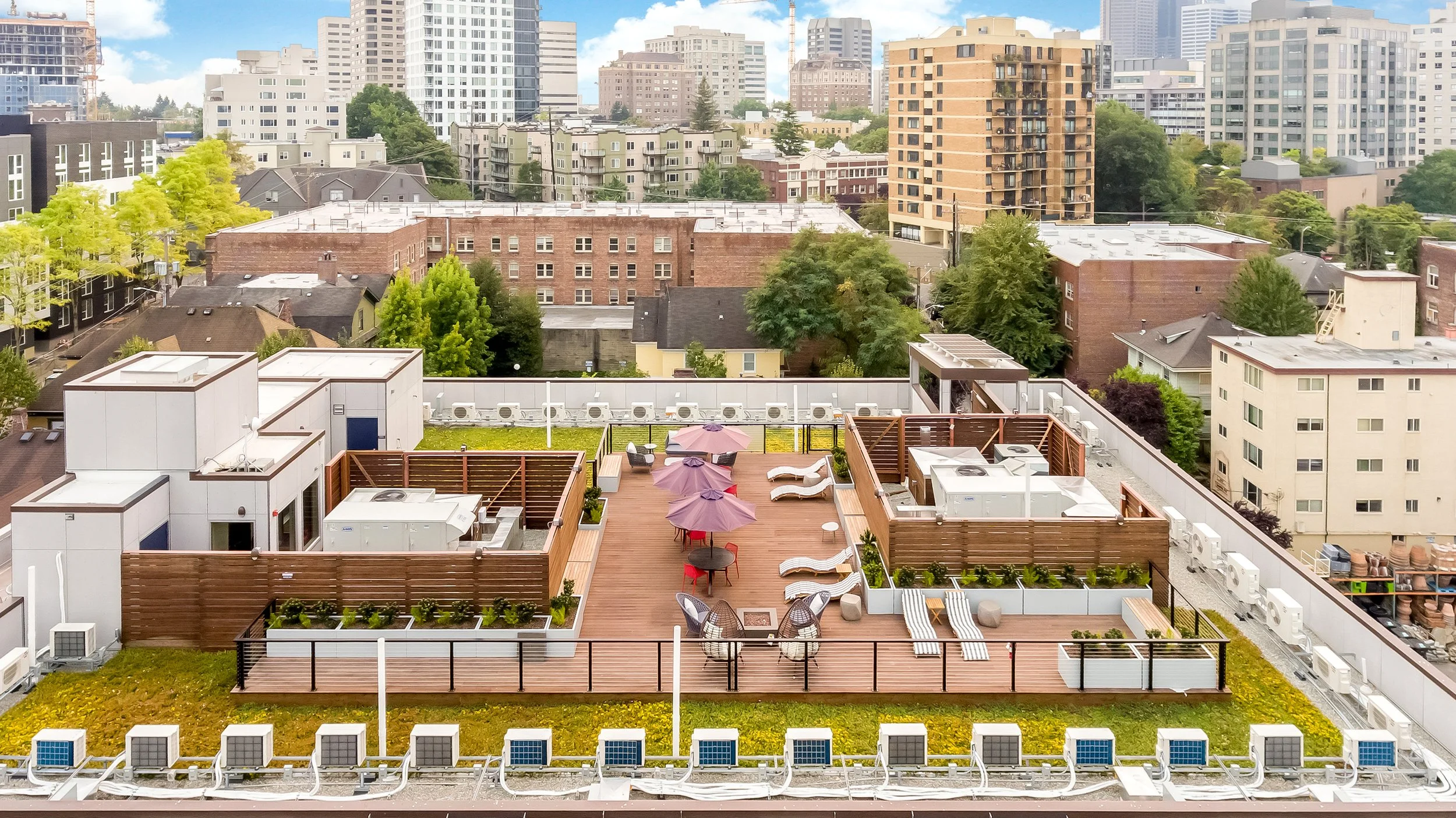 Rooftop terrace with seating, umbrellas, lounge chairs, and planters, surrounded by city buildings and trees.