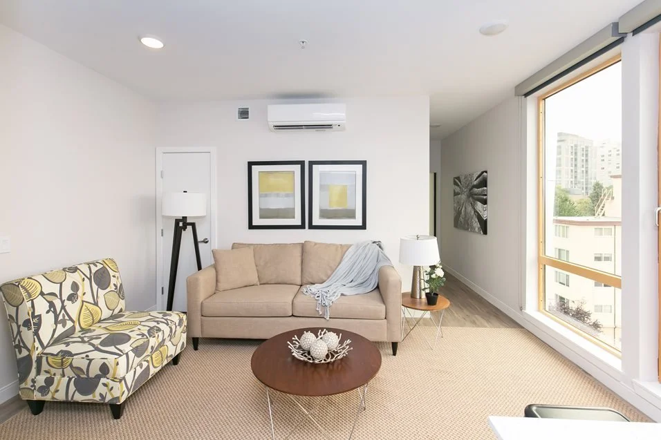 Living room with beige sofa, patterned armchair, wooden coffee table, floor lamp, table lamp, wall art, and large window with city view.