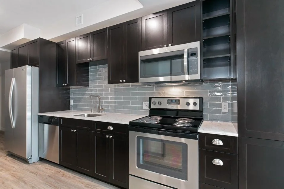 Modern kitchen with black cabinets, stainless steel refrigerator, microwave, oven, and dishwasher, gray subway tile backsplash, and white countertop.