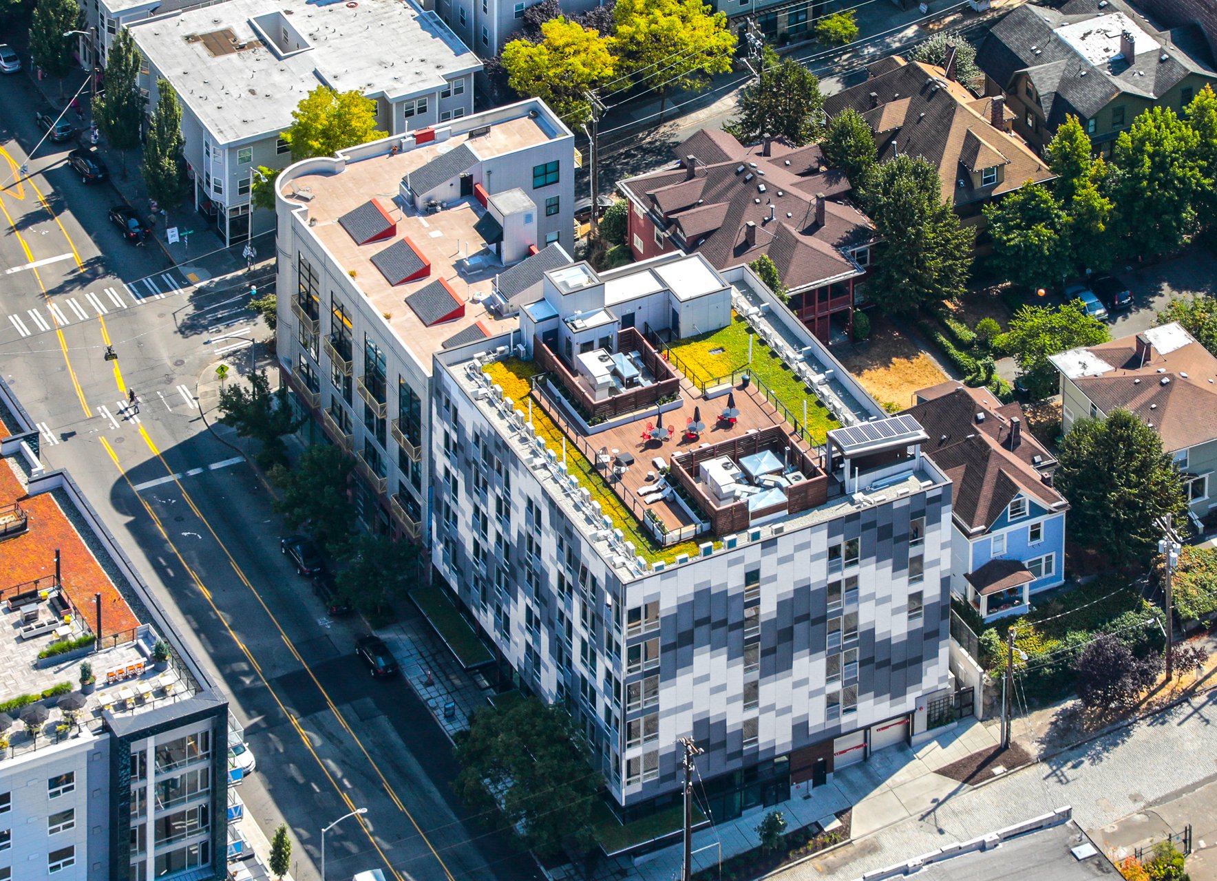 Aerial view of a modern multi-story building with a rooftop terrace, chairs, umbrellas, and greenery, surrounded by a cityscape with residential houses and streets.