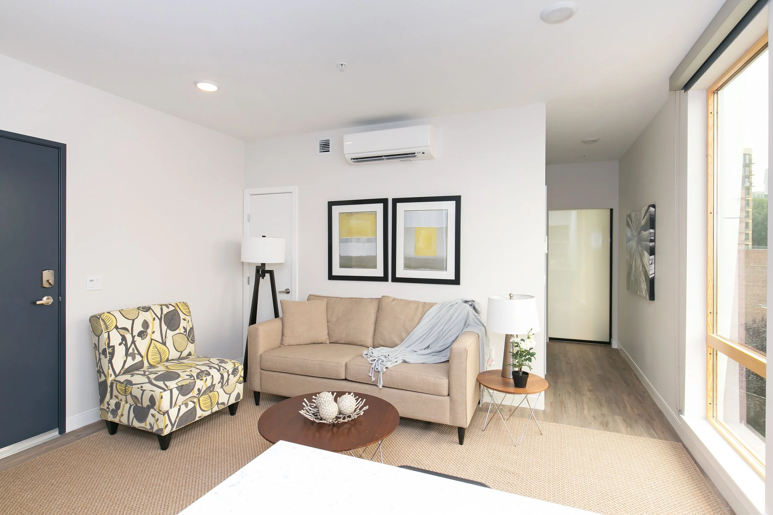 Living room with a beige sofa, a patterned armchair, a small round wooden side table with a plant, a white lamp, and framed artwork on the wall, large window on the right, and wooden flooring.