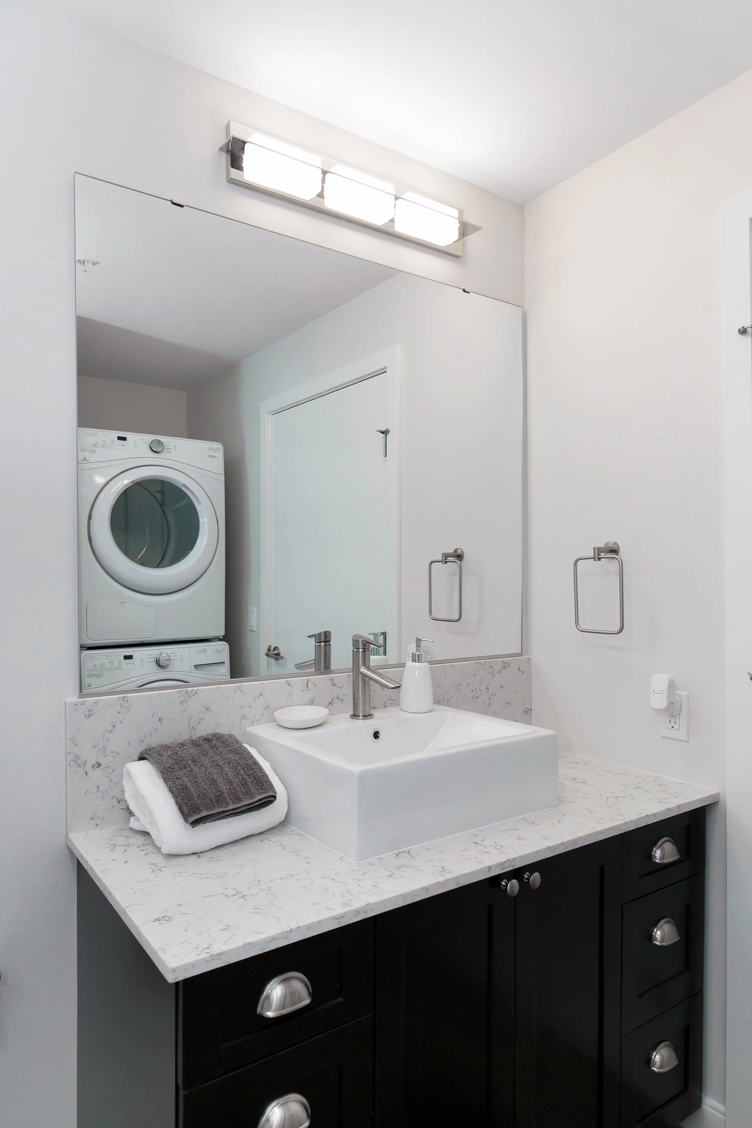 Modern bathroom with a white marble countertop, square vessel sink, and a large mirror. There are folded towels and a soap dispenser on the countertop. A stacked washer and dryer are visible in the reflection of the mirror.