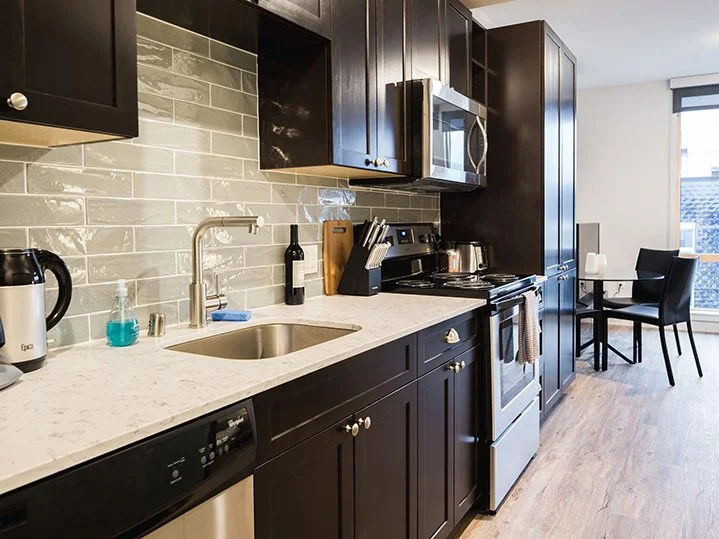Modern kitchen with dark cabinets, white marble countertop, and stainless steel appliances. Features a sink with a blue scrub brush, soap dispenser, and a bottle of wine on the counter. There's a small dining area with a black chair and table near a 