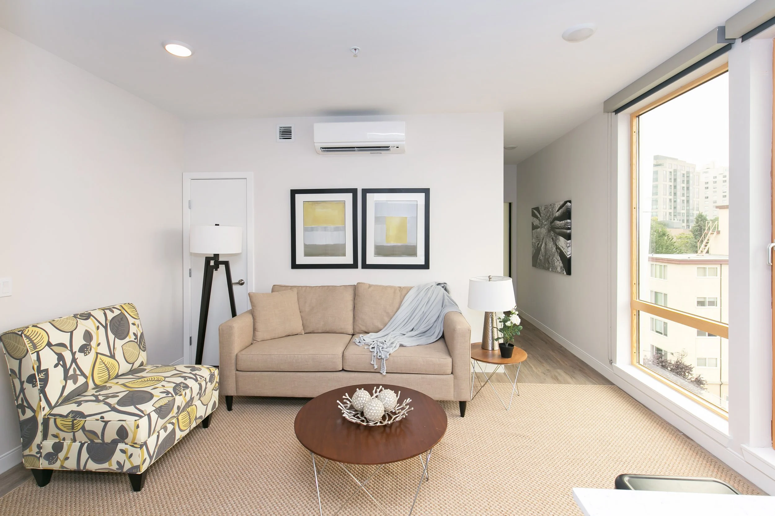 Living room with beige sofa, patterned armchair, wooden coffee table with decorative objects, side table with a lamp and plant, artwork on white walls, large window showing city buildings.
