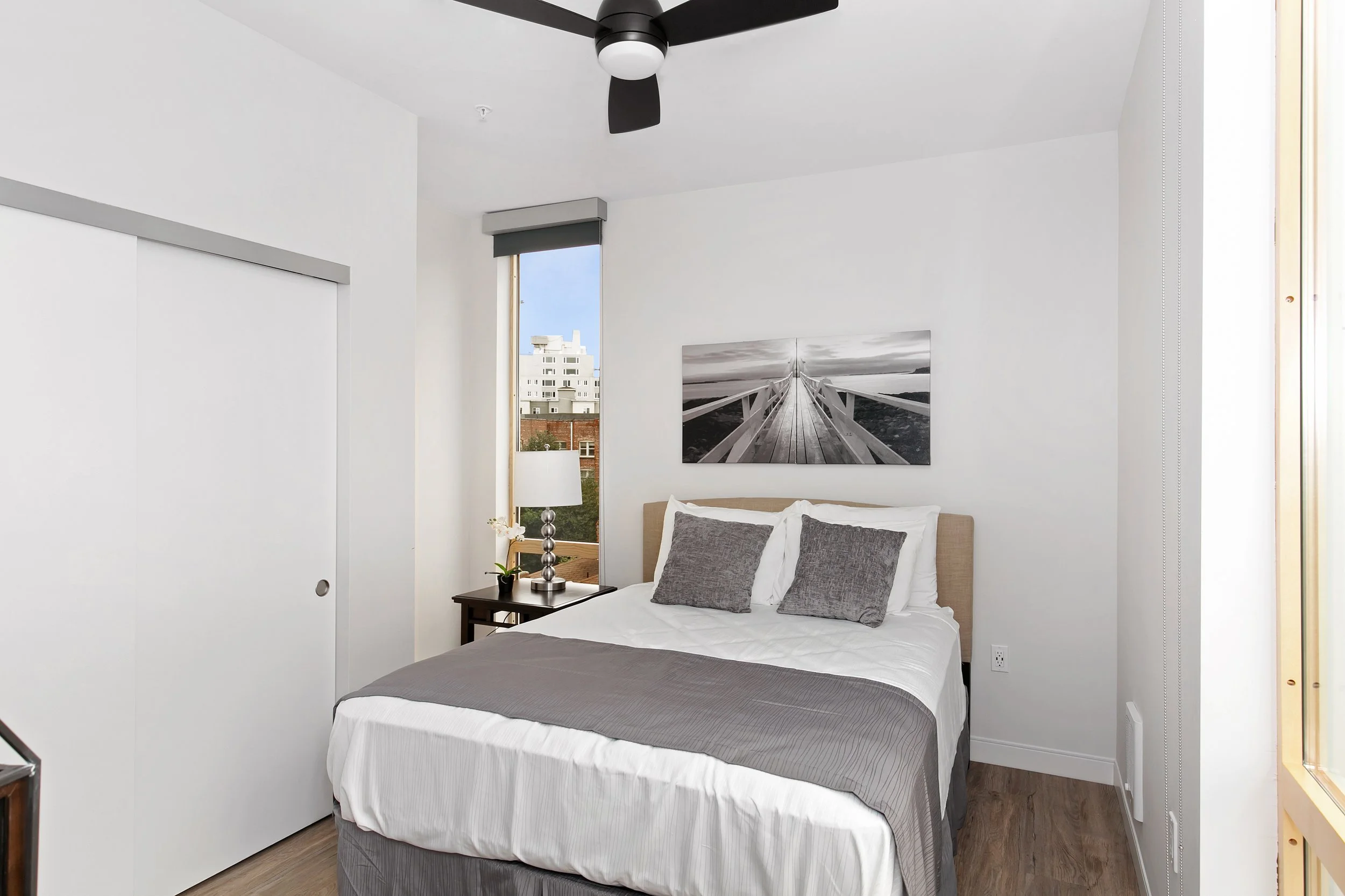 A bedroom with a bed, pillows, a window with city view, a black fan on the ceiling, a painting of a bridge above the bed, and a nightstand with a lamp and flower