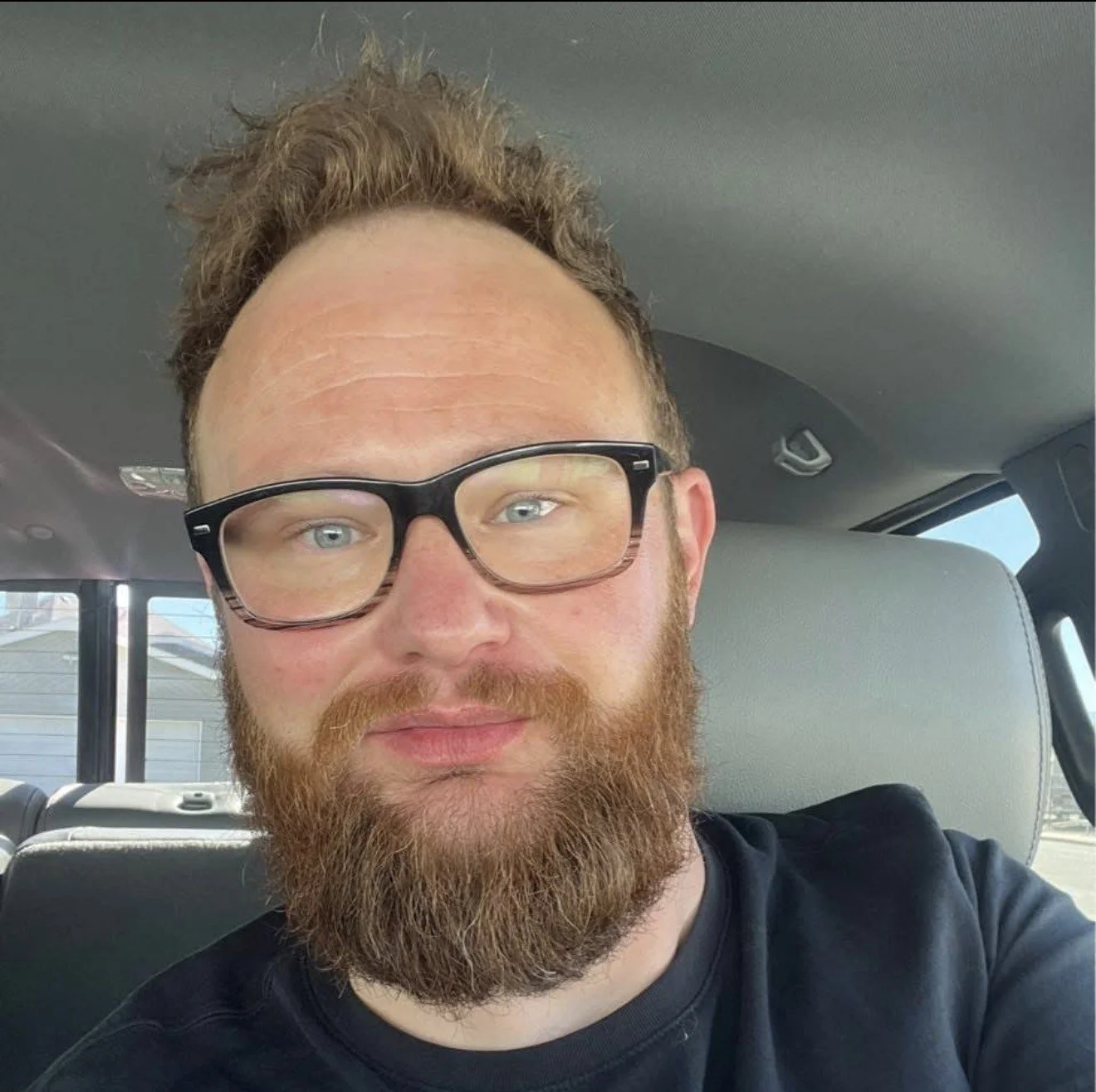 A man with glasses and a beard sitting in a vehicle, taking a selfie.
