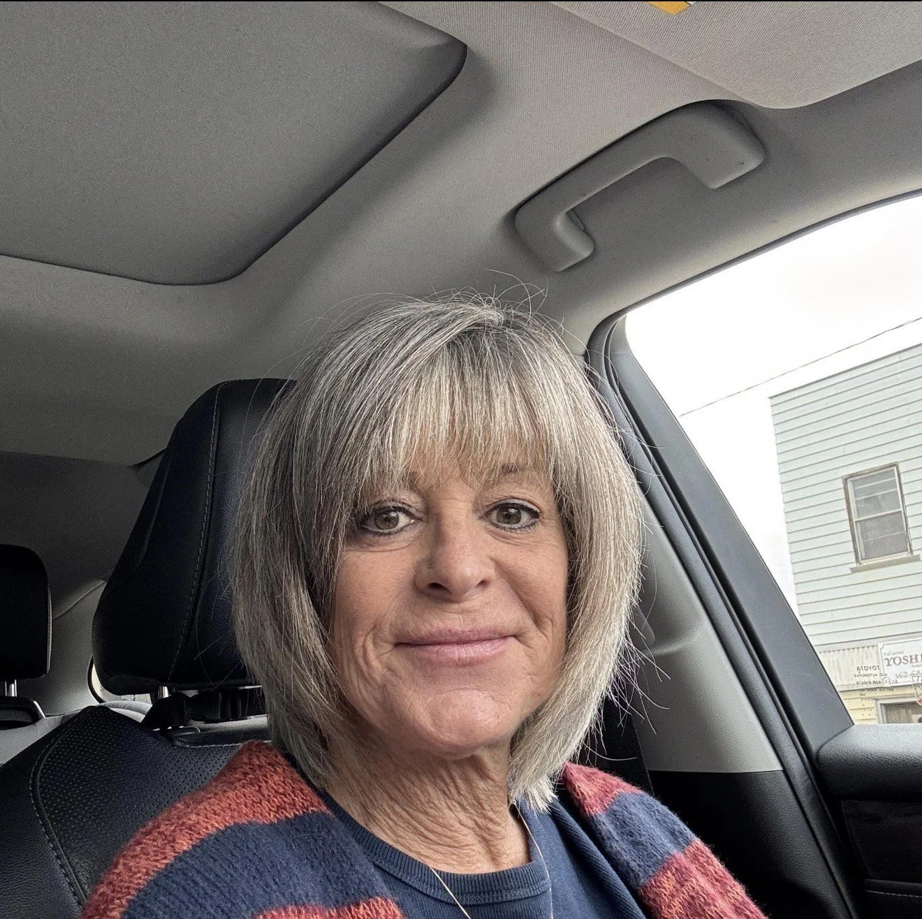 A woman with blonde hair and a striped sweater sitting in a car, looking at the camera.