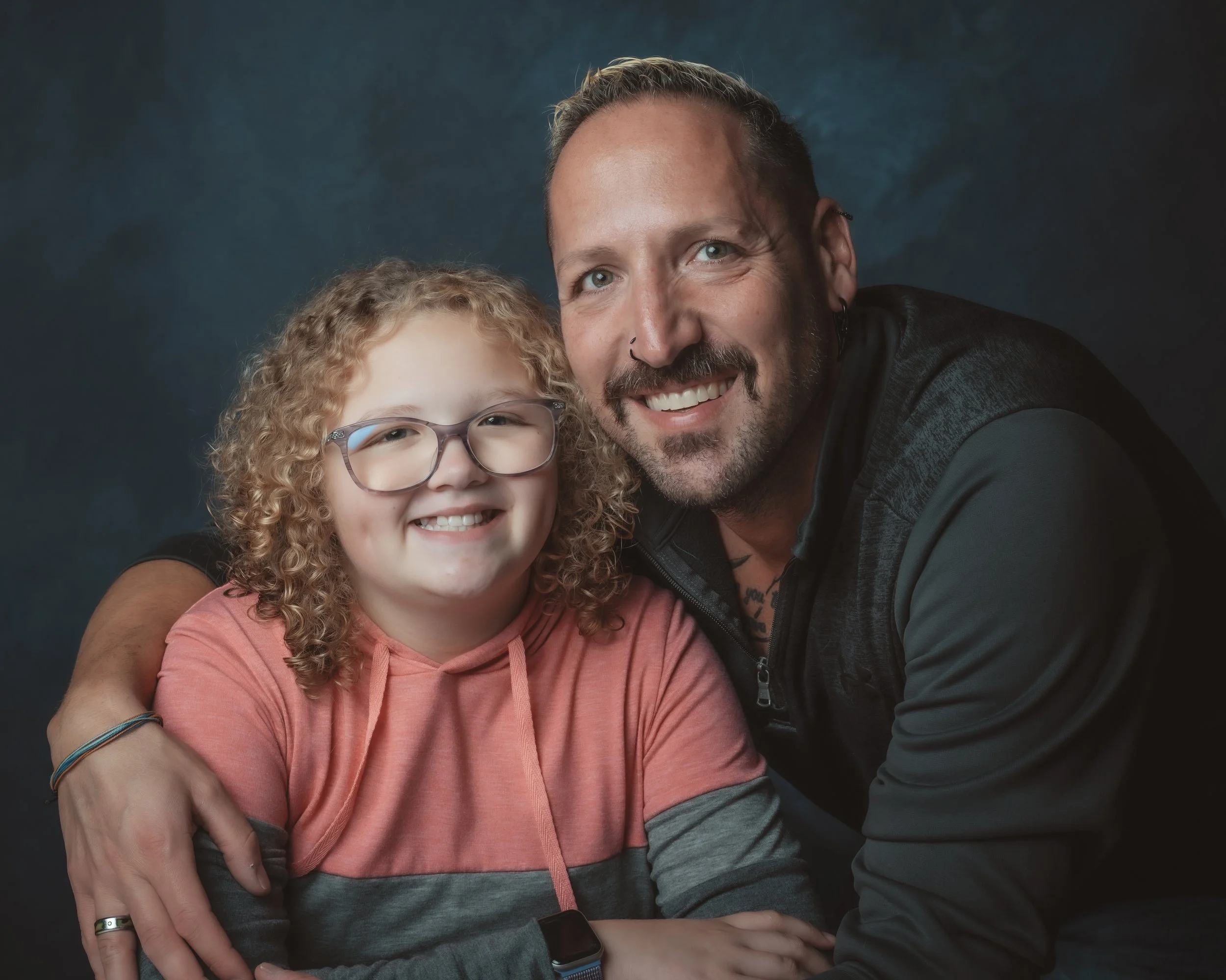A smiling man with a beard and a pierced nose, wearing a black jacket, sitting close to a young girl with curly blond hair, glasses, and a pink hoodie, against a dark background.