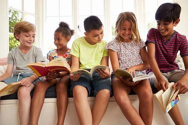 Five children sitting on a bench, reading books together, with a large window and greenery in the background.