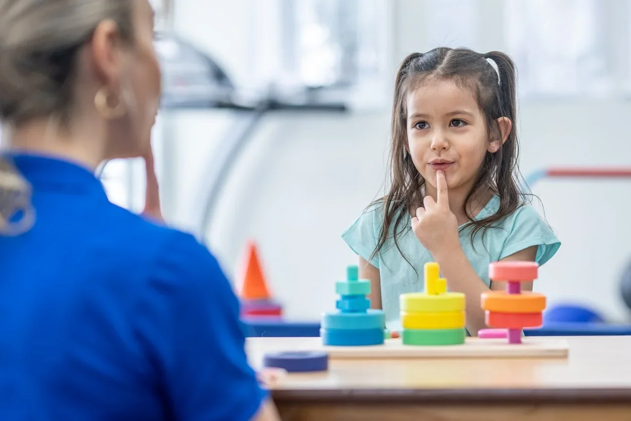 Young girl with dark hair in pigtails sitting at a table, thinking with her finger on her lip, in a clinical or therapy setting with a woman, possibly a therapist, across from her, and colorful stacking toys on the table.