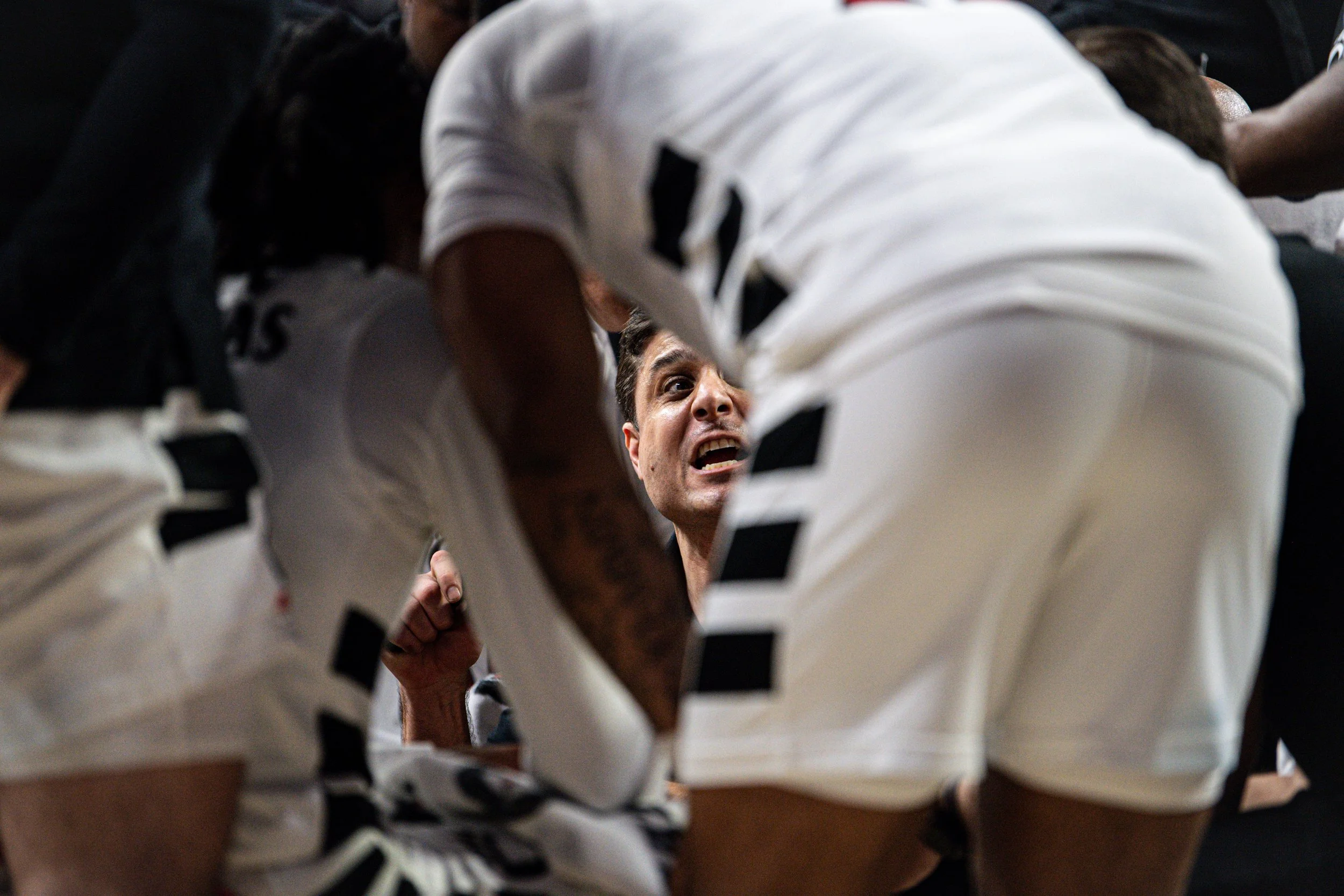 A group of athletes in a huddle, with one man in the center looking up and speaking passionately, inside a gym or sports facility.