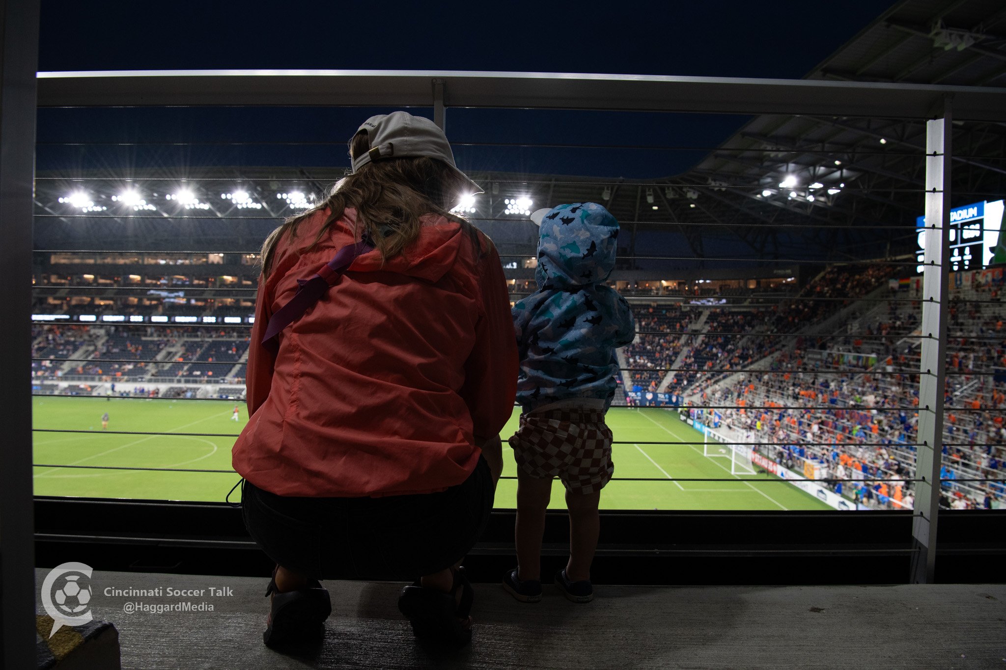 A woman and a young child watching a soccer match from a stadium balcony at night.