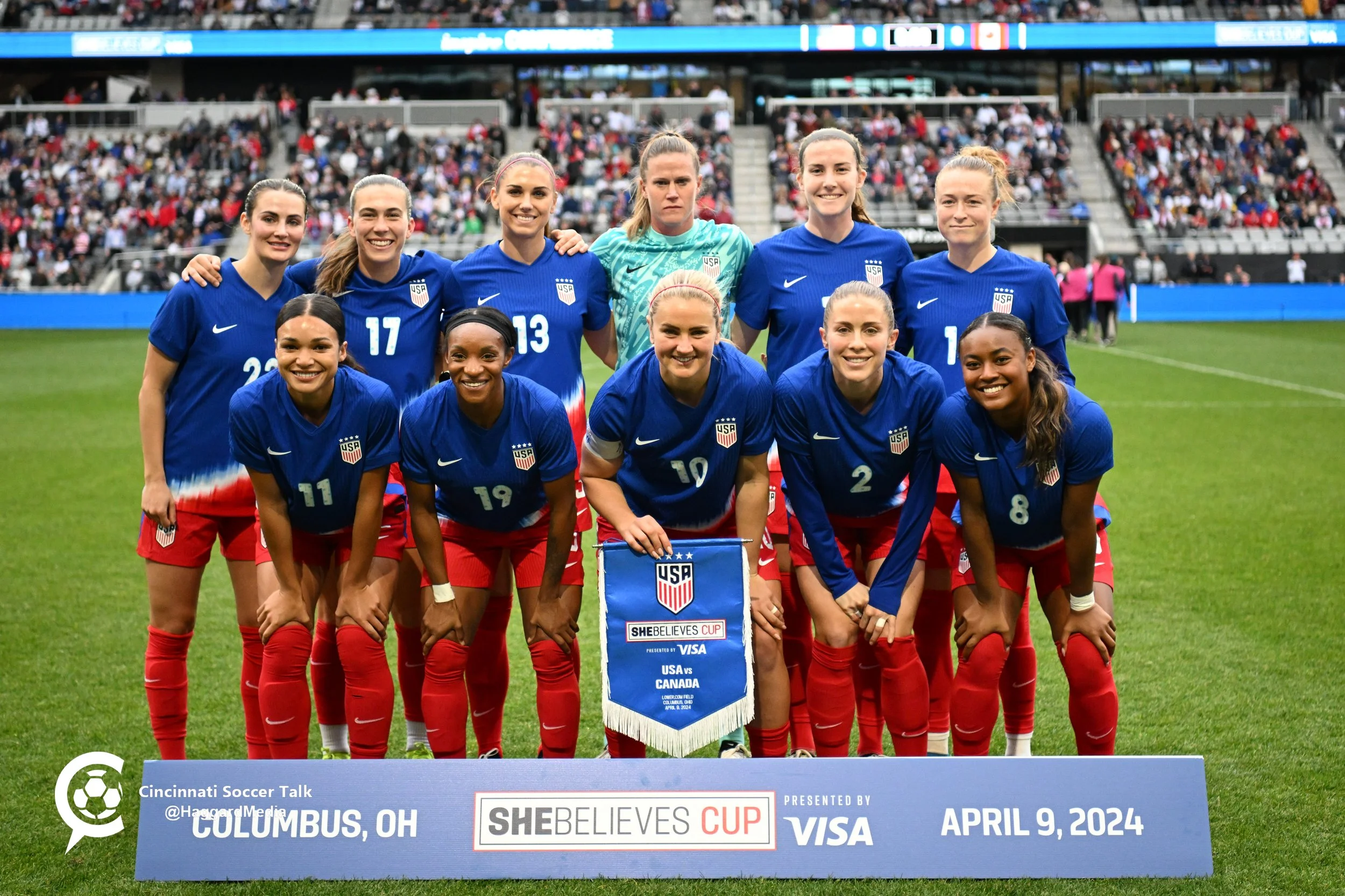 The U.S. women's national soccer team poses for a photo on the field before a match, wearing blue and red uniforms, holding a SheBelieves Cup banner, with a crowd in the stadium stands behind them, dated April 9, 2024, in Columbus, Ohio.
