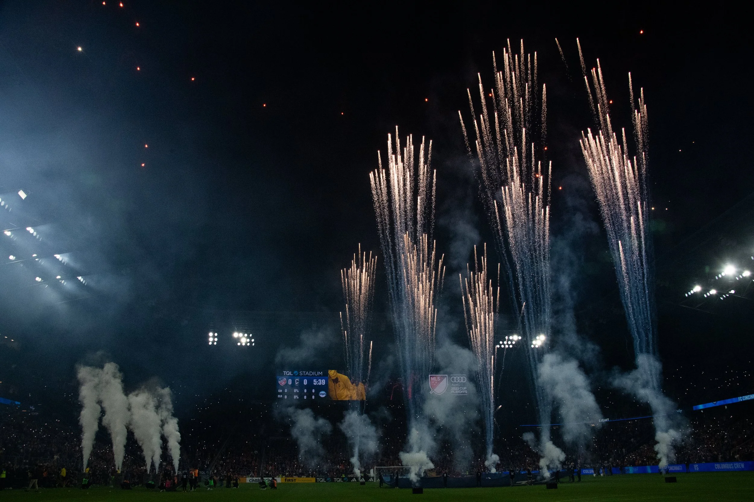 Fireworks display over a stadium during a night event with bright streaks of fireworks and smoke.