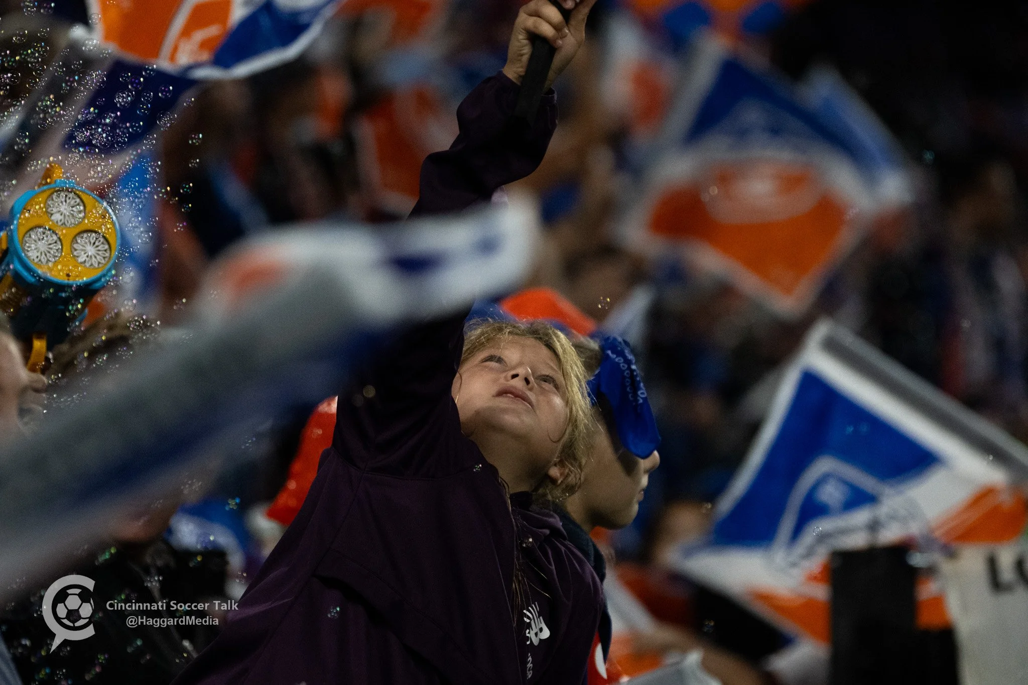A young woman in a black jacket raising her arm at a soccer game surrounded by fans with orange and blue flags and banners.