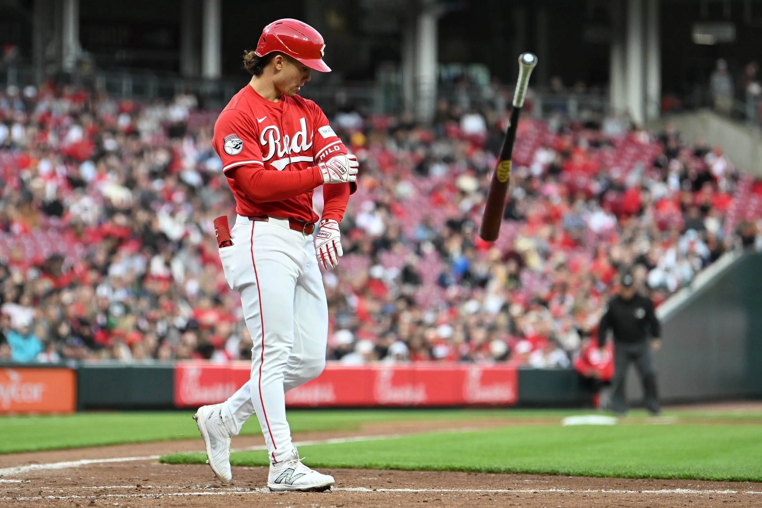 A baseball player in a red and white uniform standing on a baseball field during a game, with a crowd in the stands behind him.