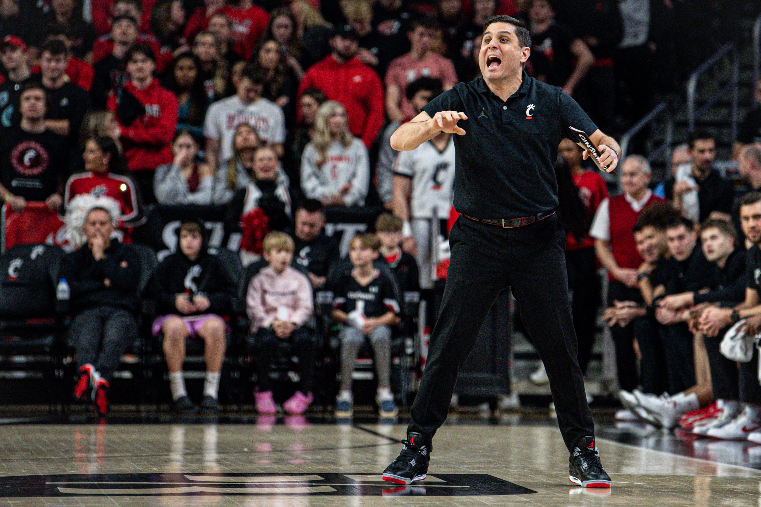 A man coach or speaker passionately gesturing in front of a crowd of children and adults, all wearing sports apparel, during a sports event at an indoor basketball court.