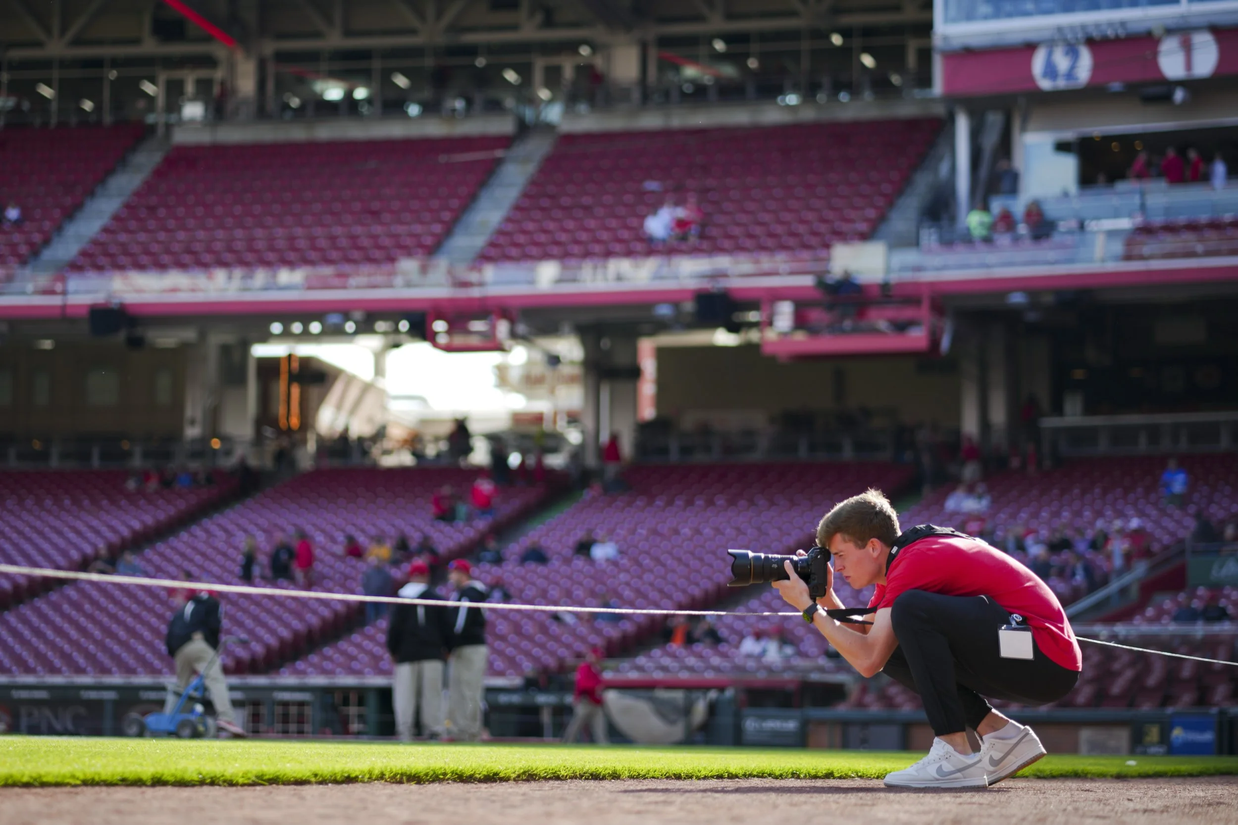 Person crouching and taking a photo with a camera at a sports stadium, with empty red seats and some people in the background.